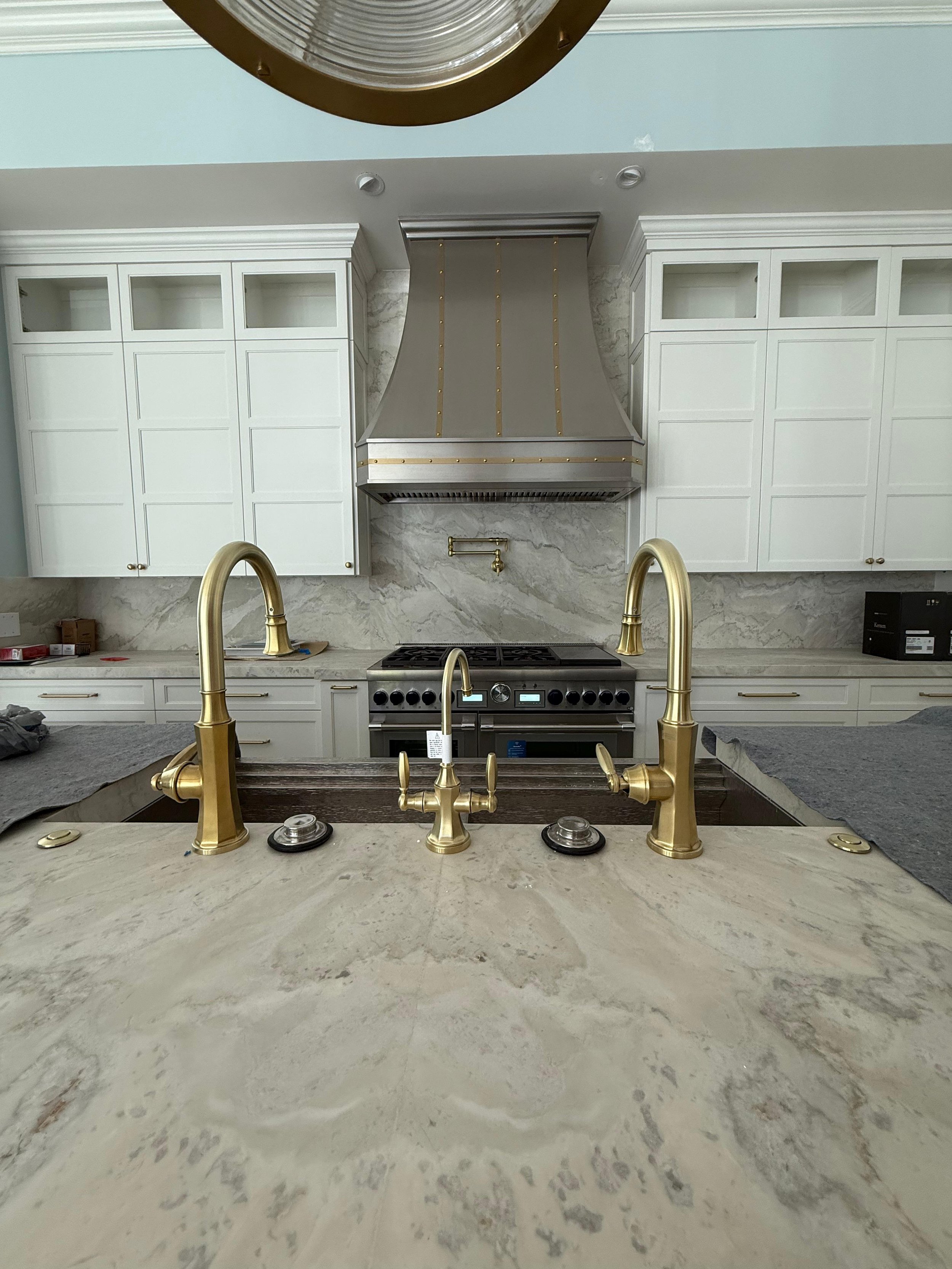 Kitchen with a marble countertop, gold-colored dual faucets, a stove with stainless steel range, white upper and lower cabinets, and a large stainless steel range hood.