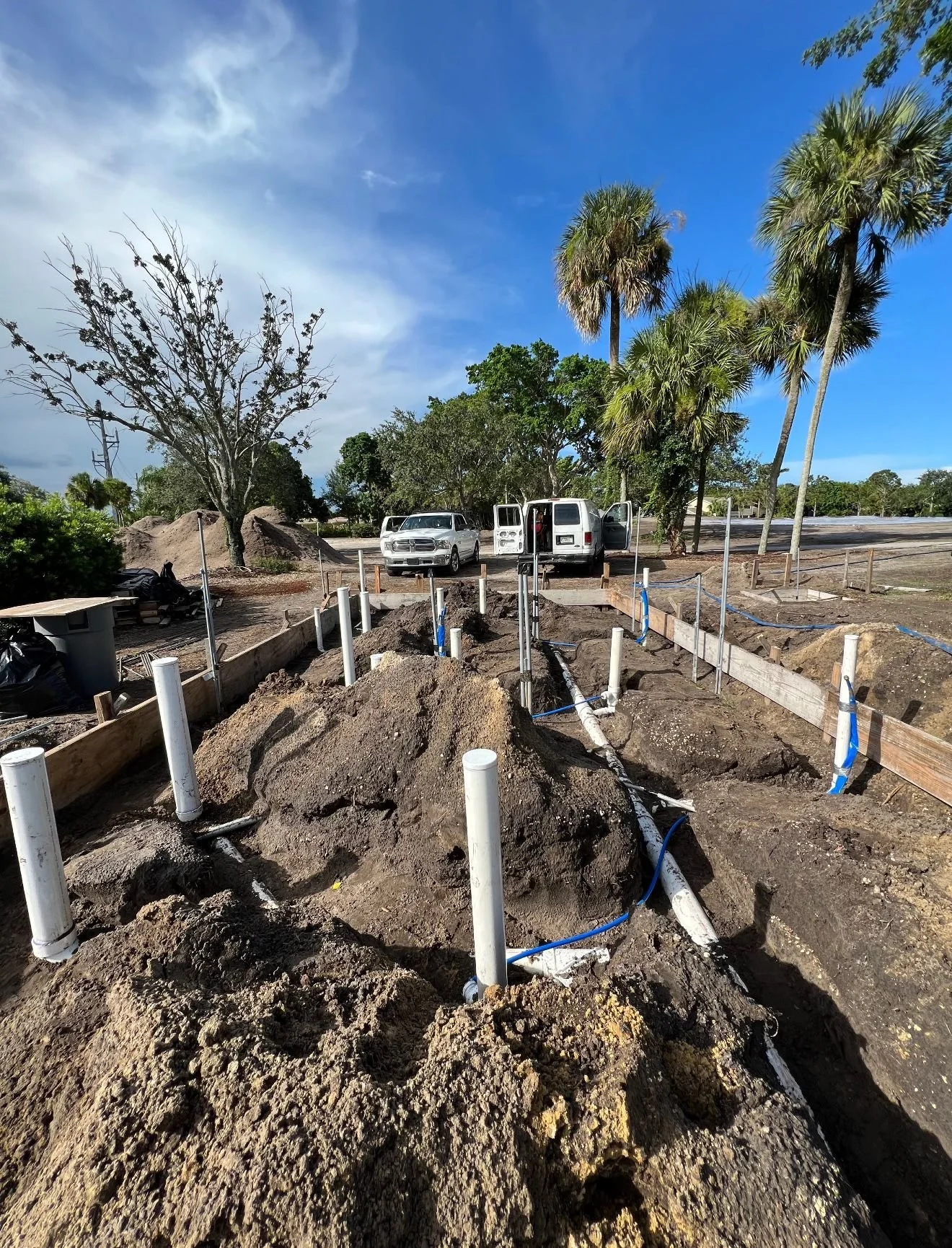Construction site with exposed pipes and dirt, surrounded by wooden barriers, with parked vehicles and palm trees in the background under a partly cloudy sky.