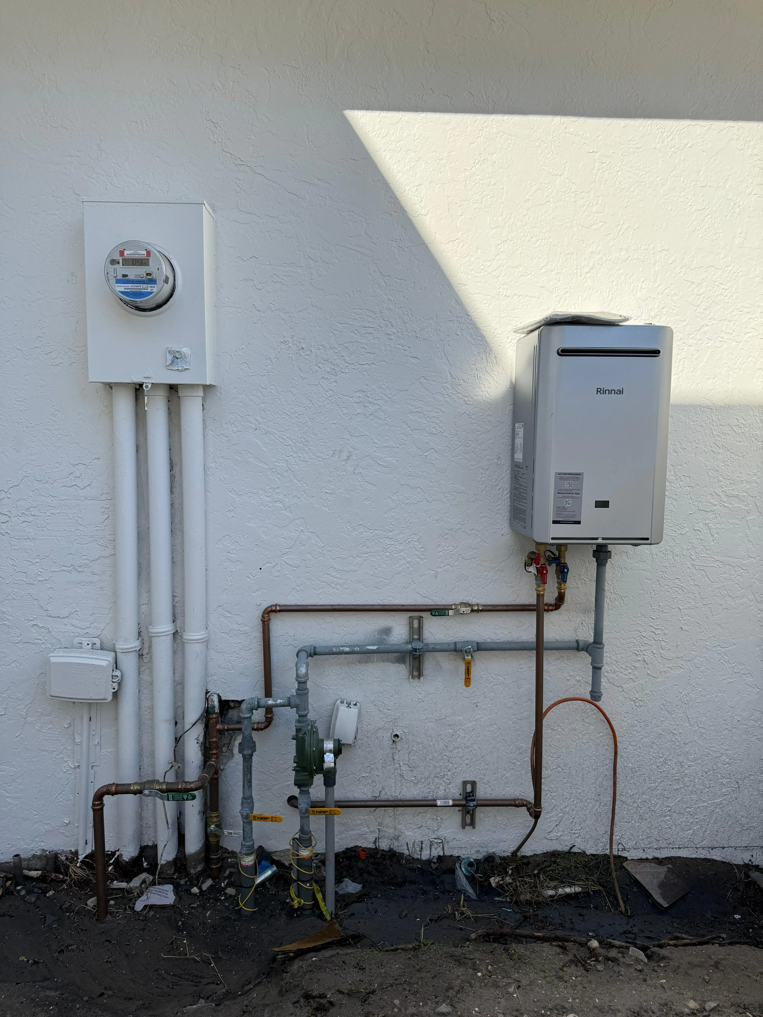 Outdoor view of utility equipment including a white electrical meter box, pipes, and a Rinnai tankless water heater mounted on a white textured wall.