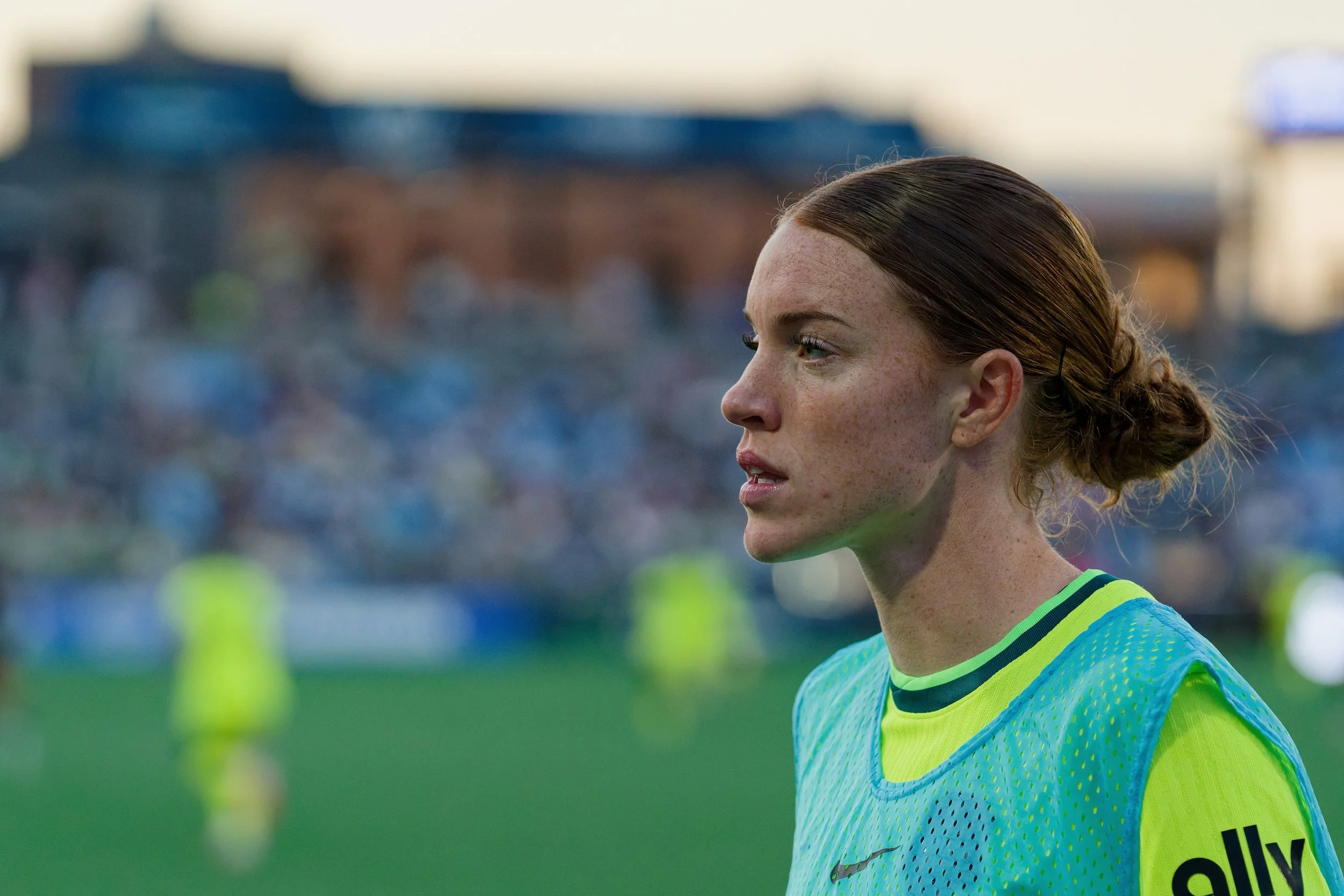 Close-up of a female soccer player with auburn hair tied in a bun, wearing a yellow and blue uniform, standing on a soccer field.