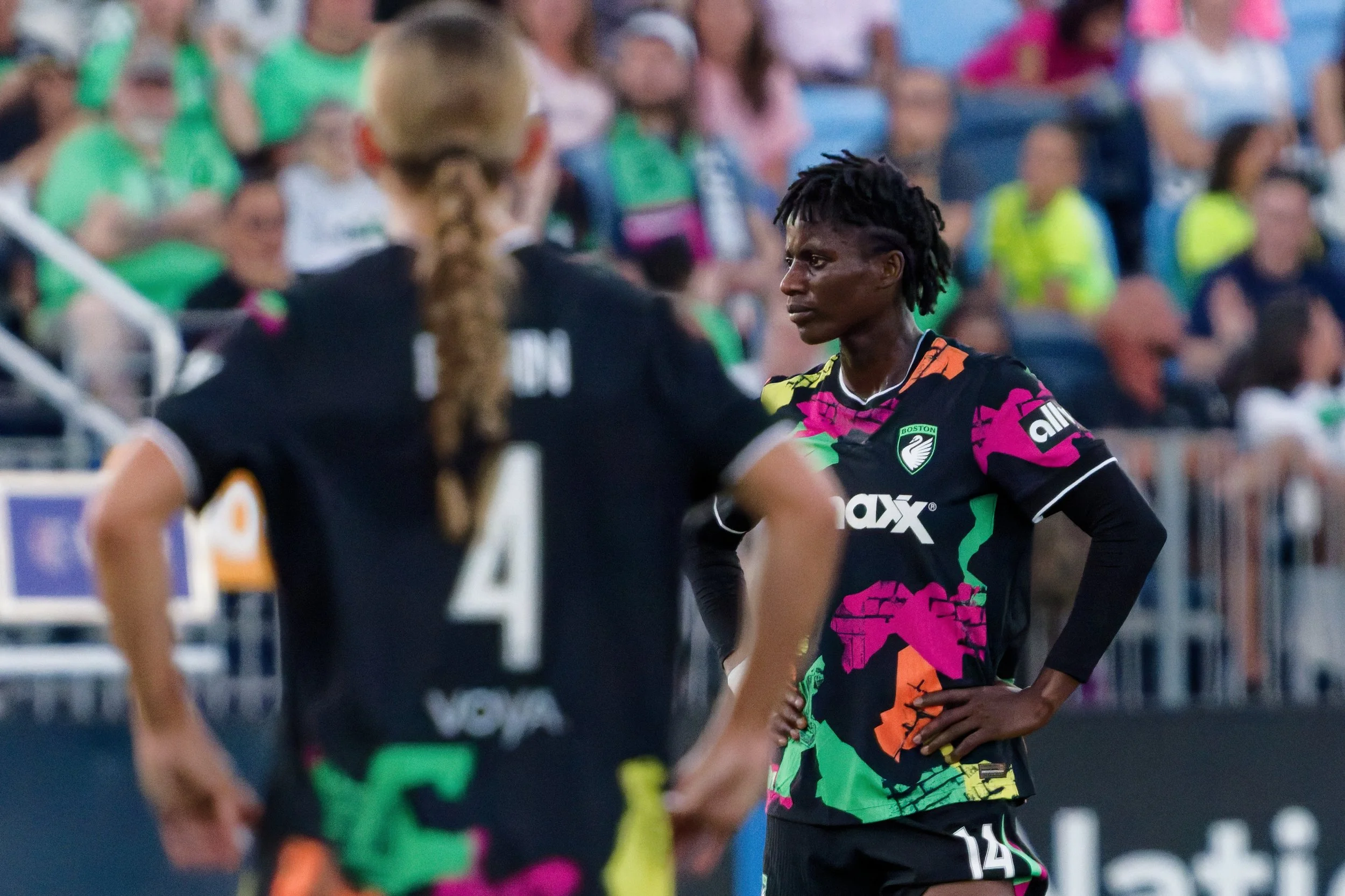 A female soccer player with dark skin and dreadlocks, wearing a colorful black, pink, green, and orange uniform with the Boston logo, standing with her hand on her hip during a game. There are other players and spectators in the background.
