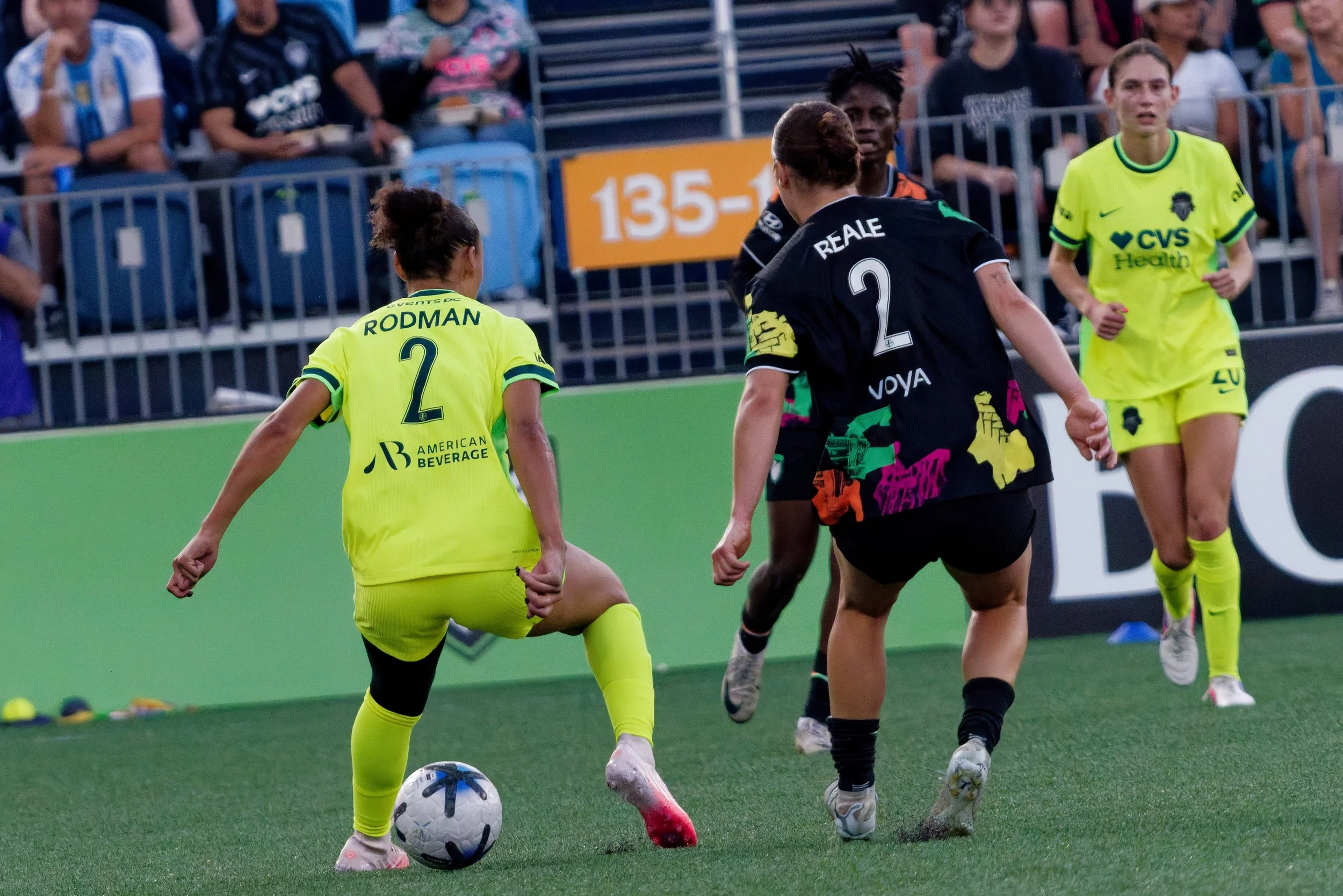 Women soccer players on the field, one in a yellow uniform with number 2, controlling the ball, while others compete for the ball during a match, with spectators in the background.