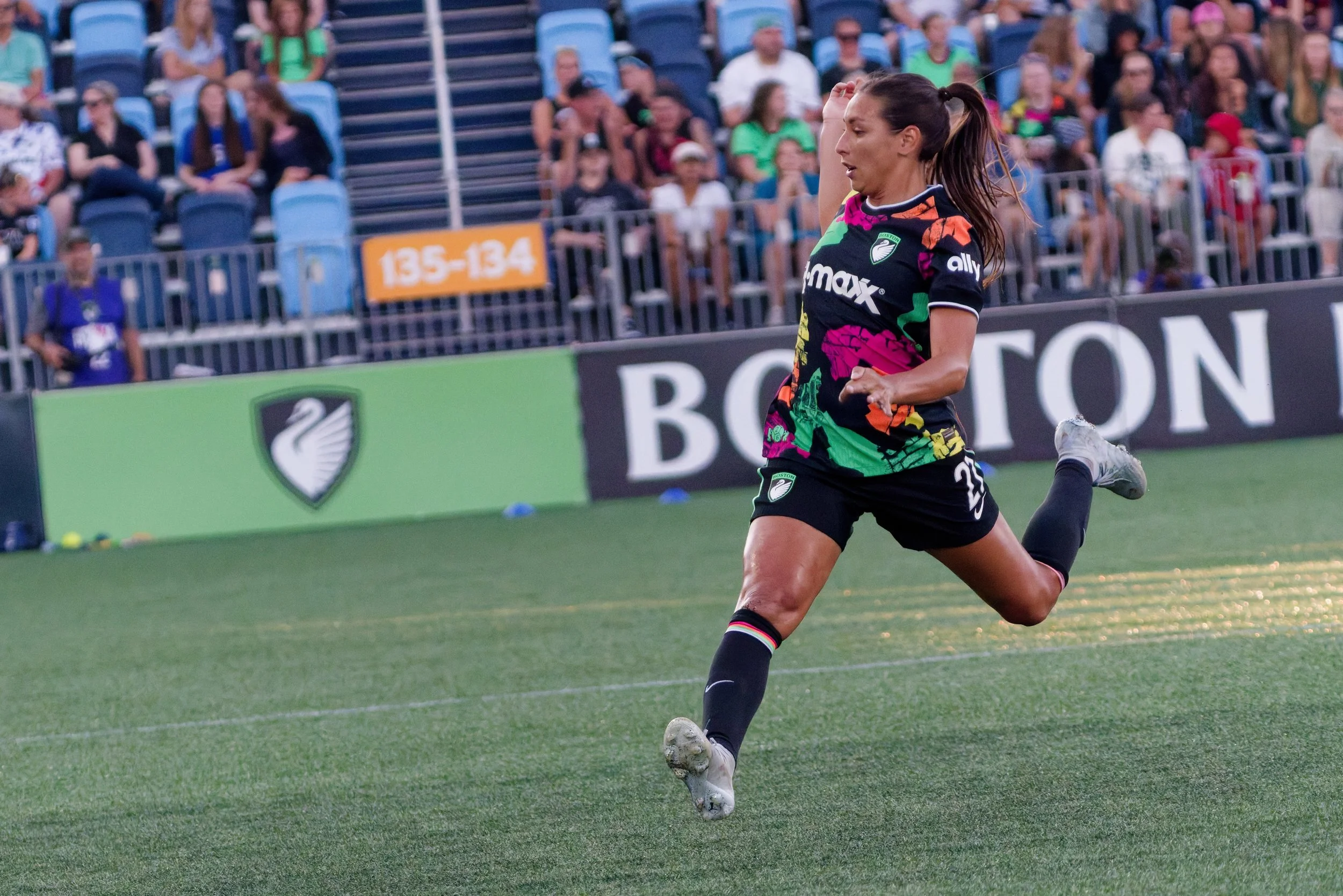A female soccer player in a colorful uniform running on the field during a game with spectators in the stands.
