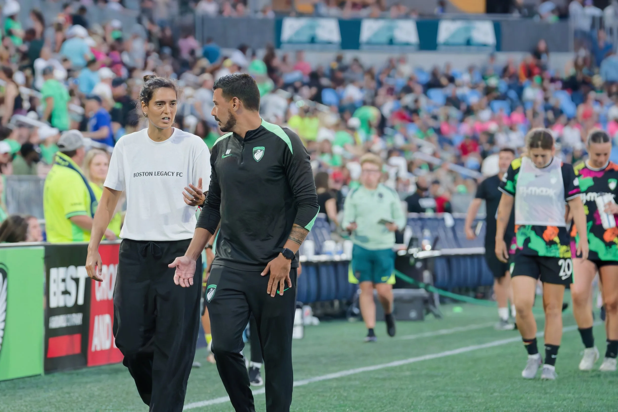 Two soccer coaches engaged in a conversation on the sideline during a match with a crowd in the stands behind them.