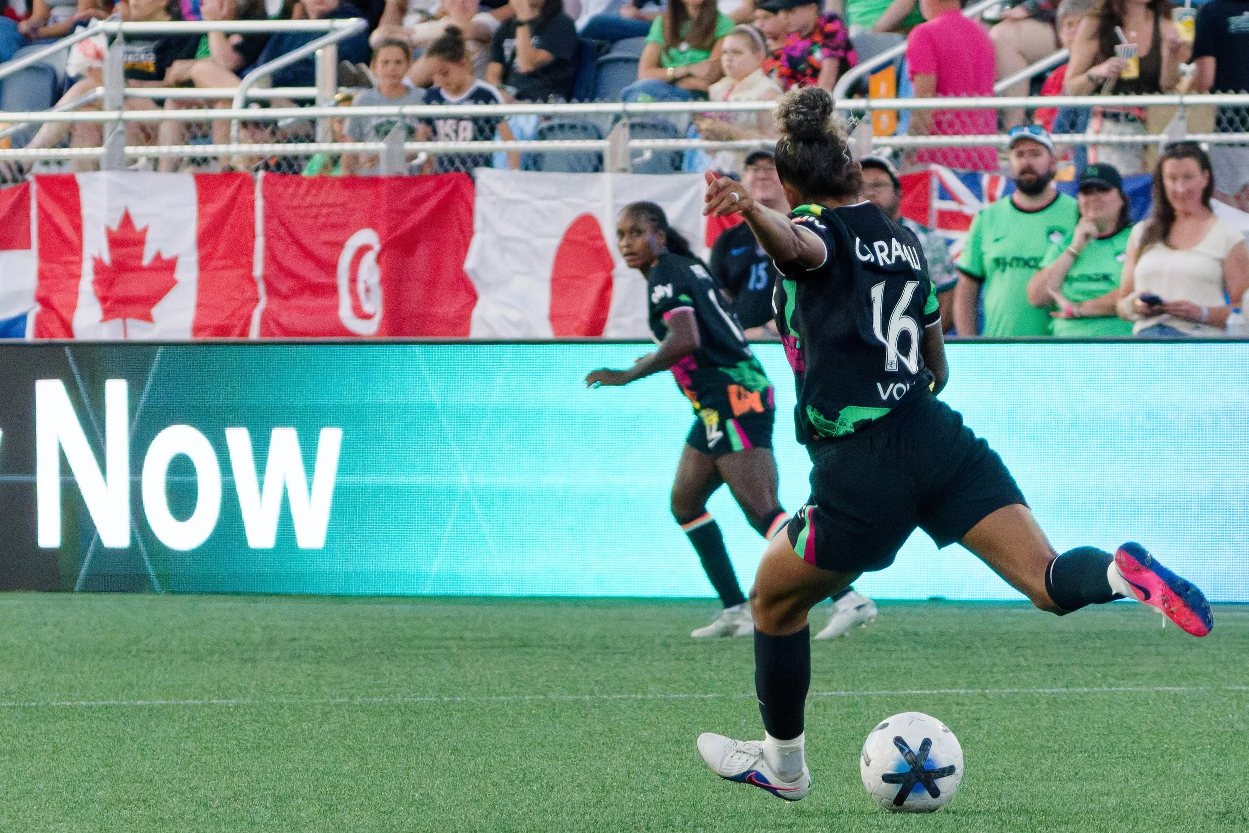 A soccer player in black shorts and shoes is in action on the field with a soccer ball at her feet, wearing a jersey with the name 'Craw' and number 16. In the background, some fans are watching the game, with volleyballs hanging over the fence and C