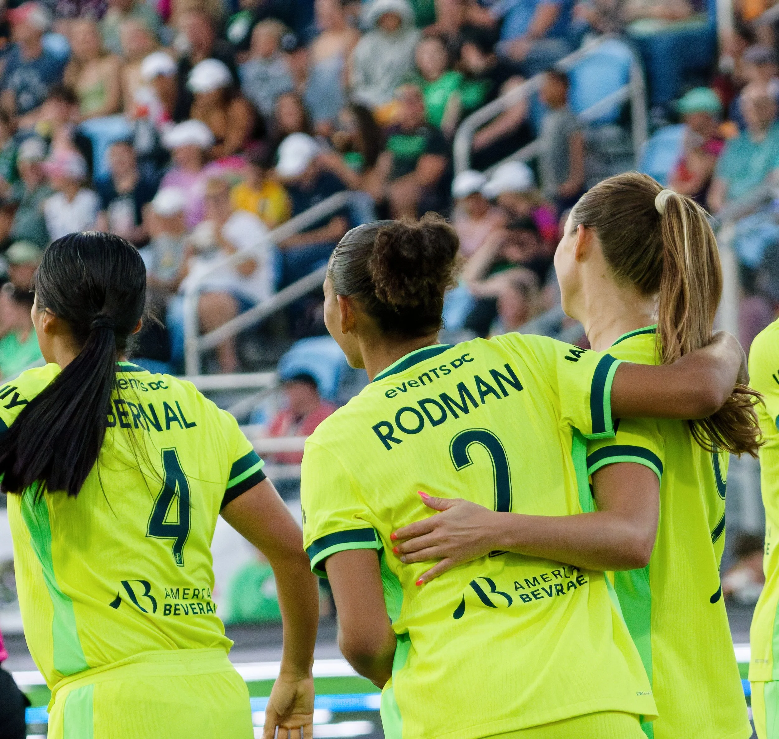 Group of female soccer players hugging during a game, wearing bright yellow jerseys with green accents, with a blurred crowd in the background.