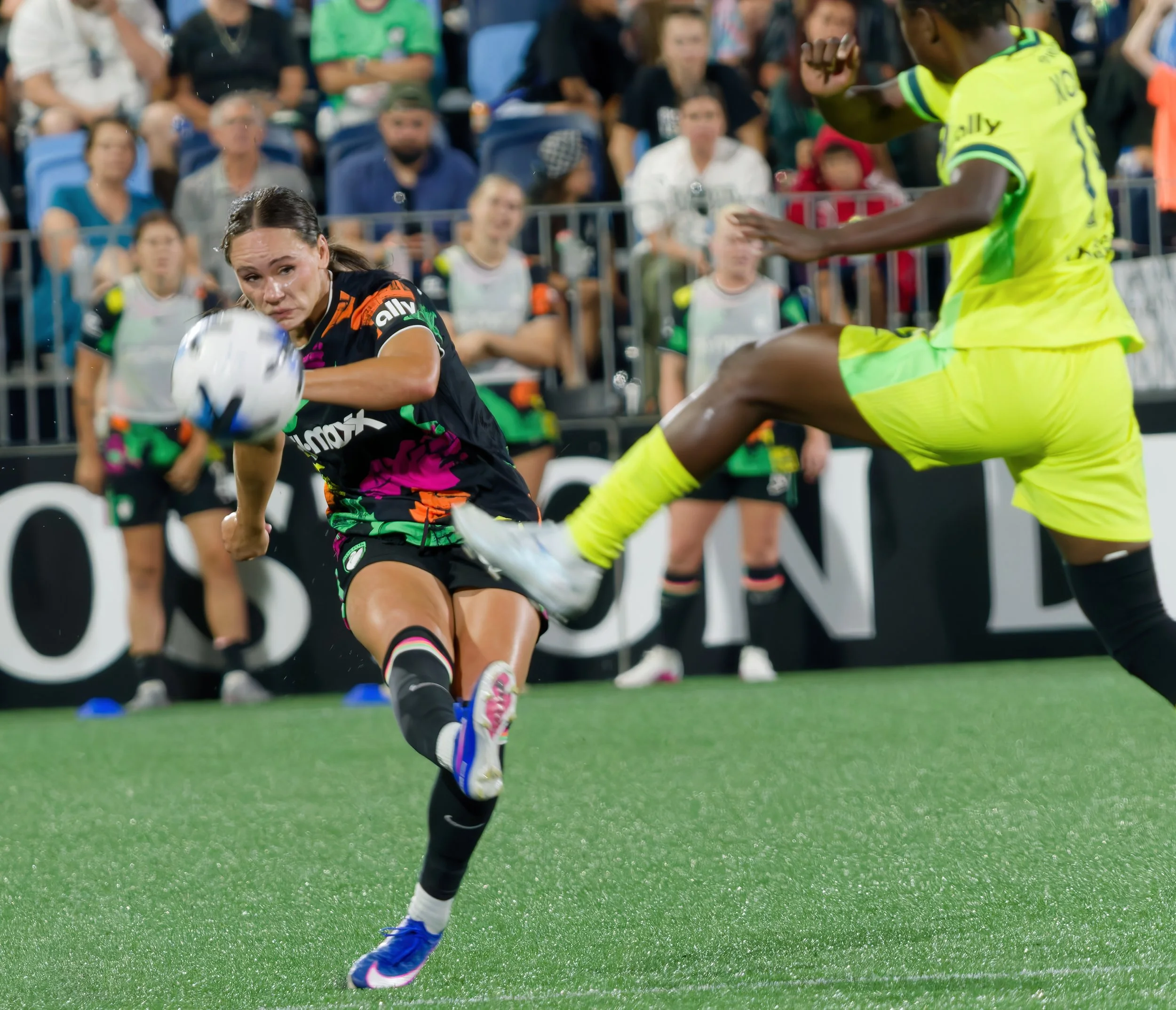 A female soccer player in a colorful jersey lunges to kick a soccer ball during a match, with spectators watching in the background.