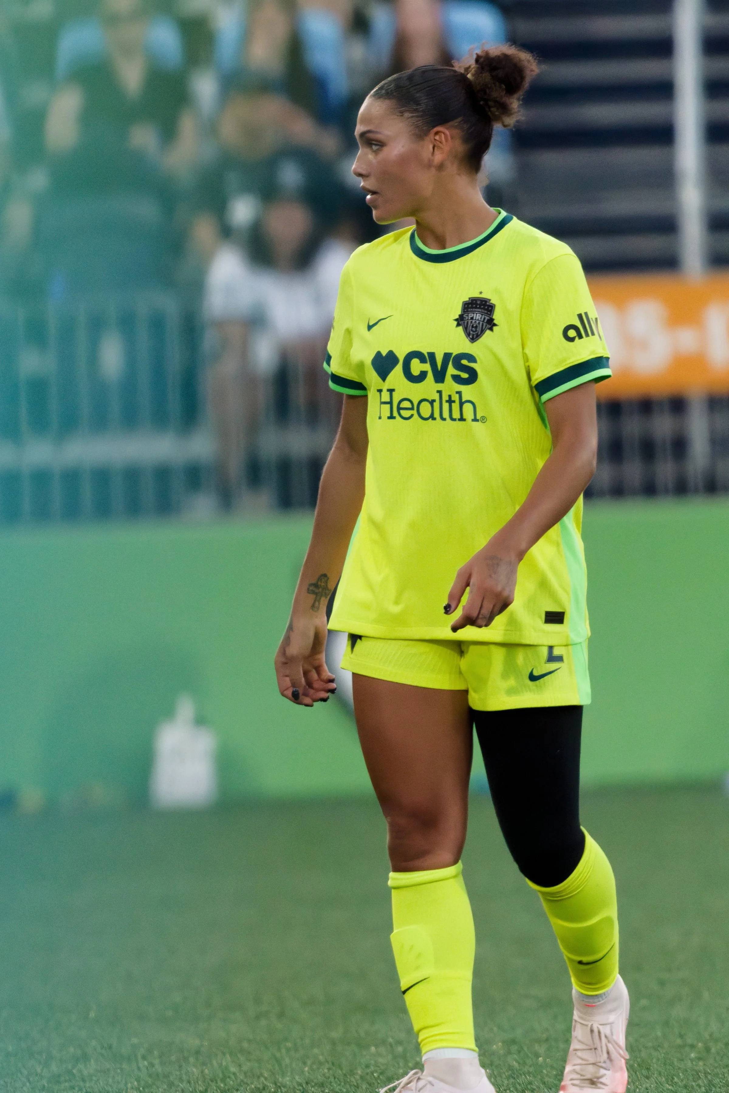 A female soccer player in yellow and black uniform on the field, with crowd and stadium in the background.