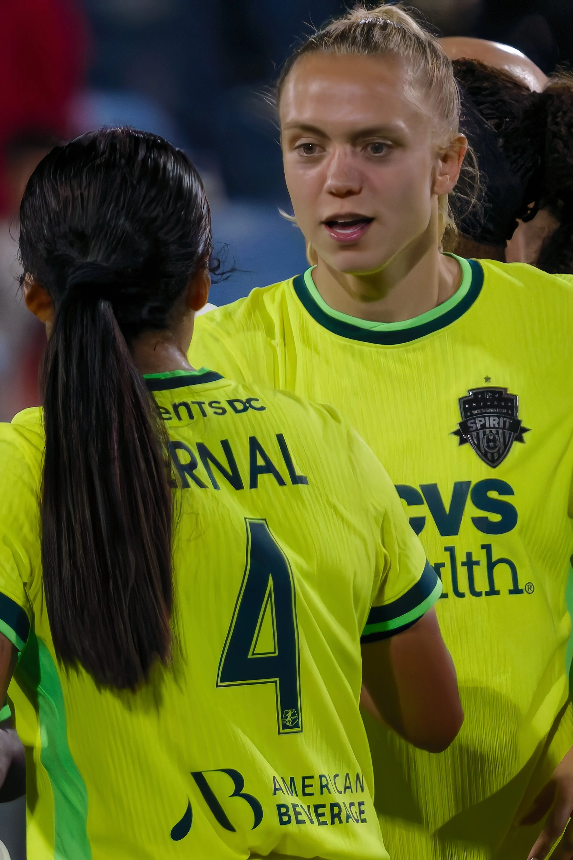 Two women wearing bright yellow soccer uniforms with black and green accents, communicating on a sports field.
