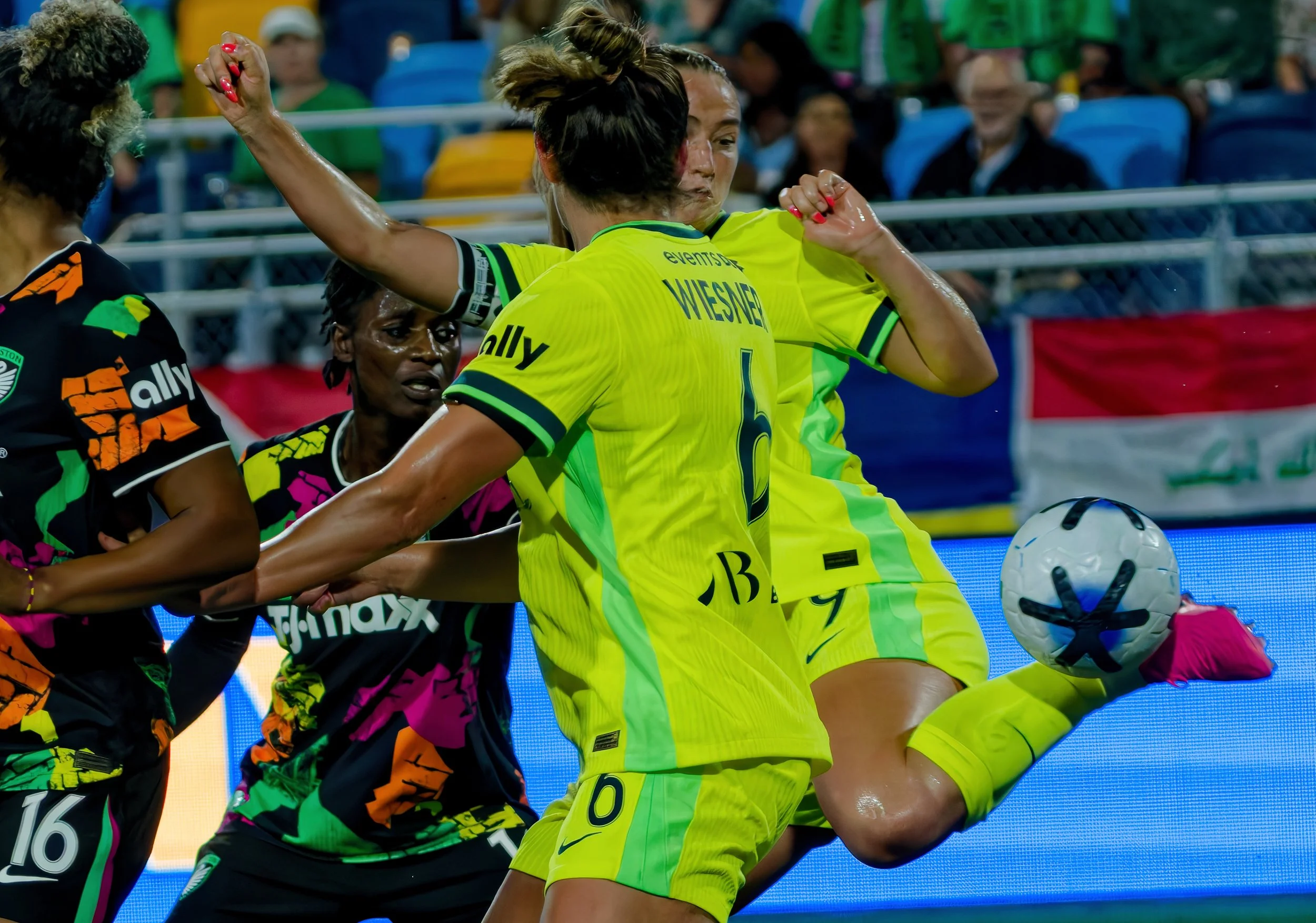 Soccer players competing for the ball during a match, with one player in a yellow uniform and other players in colorful black uniforms.