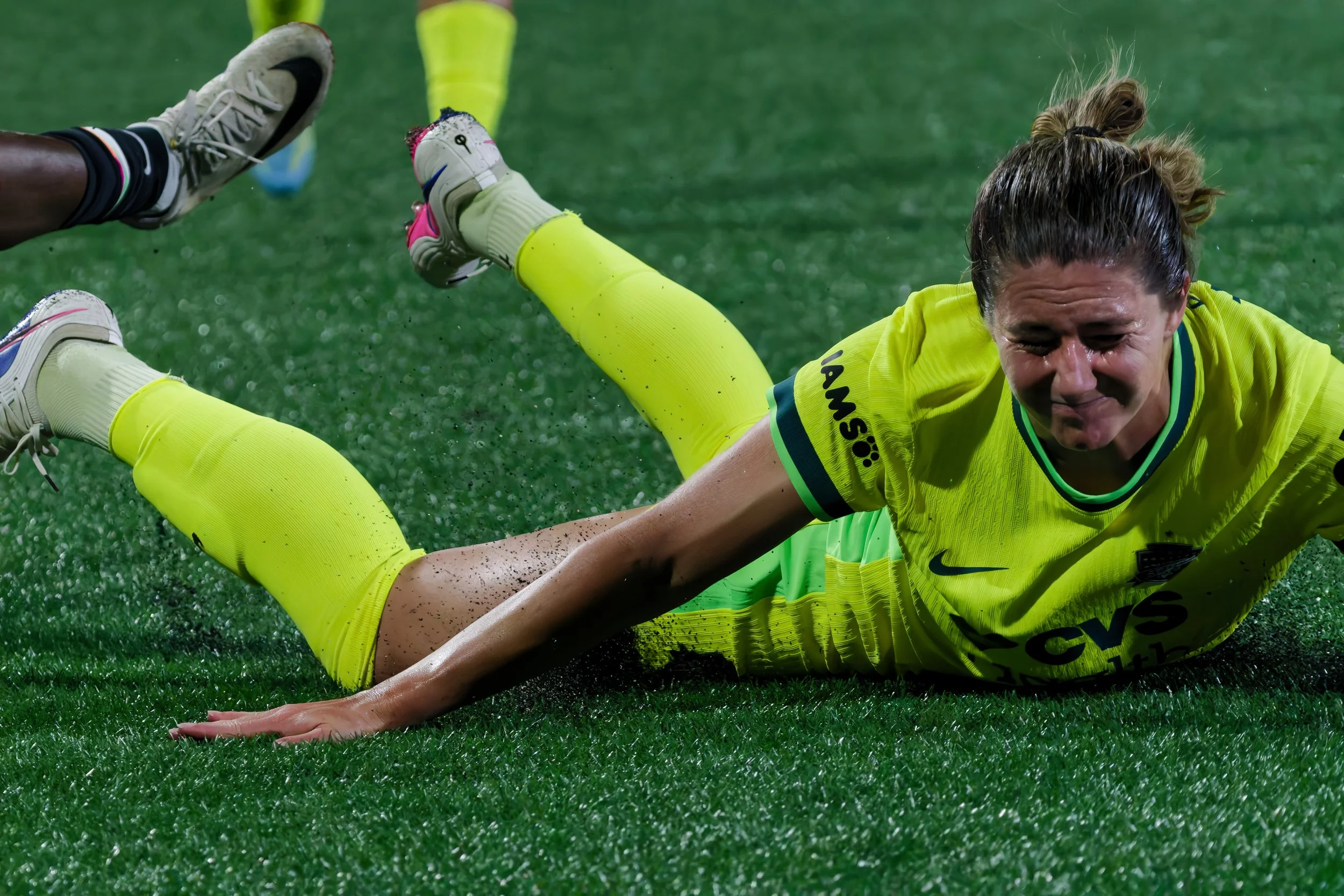 A female soccer player in yellow uniform on the field, lying on her side, with her face showing intense effort, on a wet, grassy field.
