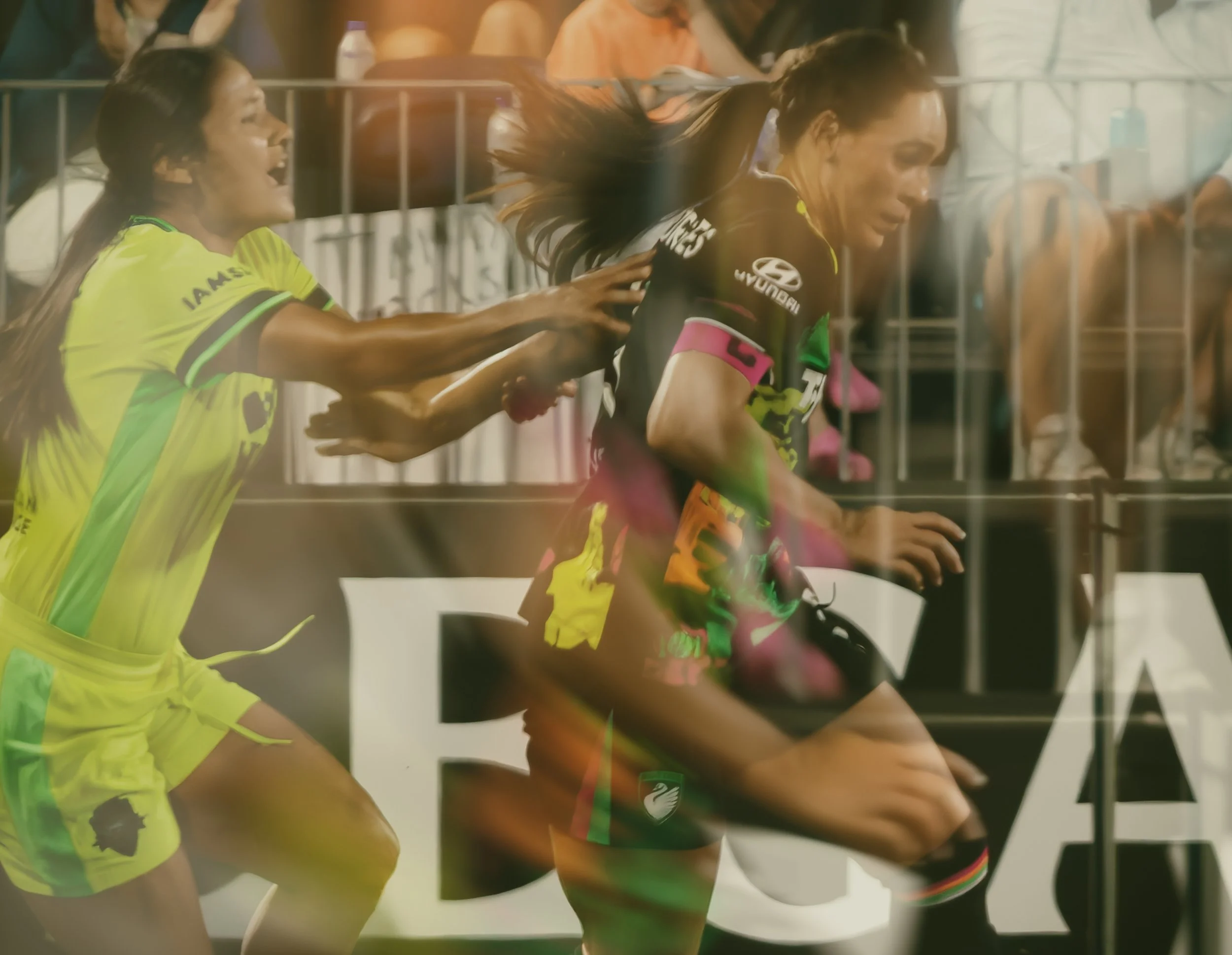 Two female athletes in black and colorful sports uniforms running during a race, with a woman in a neon yellow shirt encouraging them from the side, all behind a glass window.