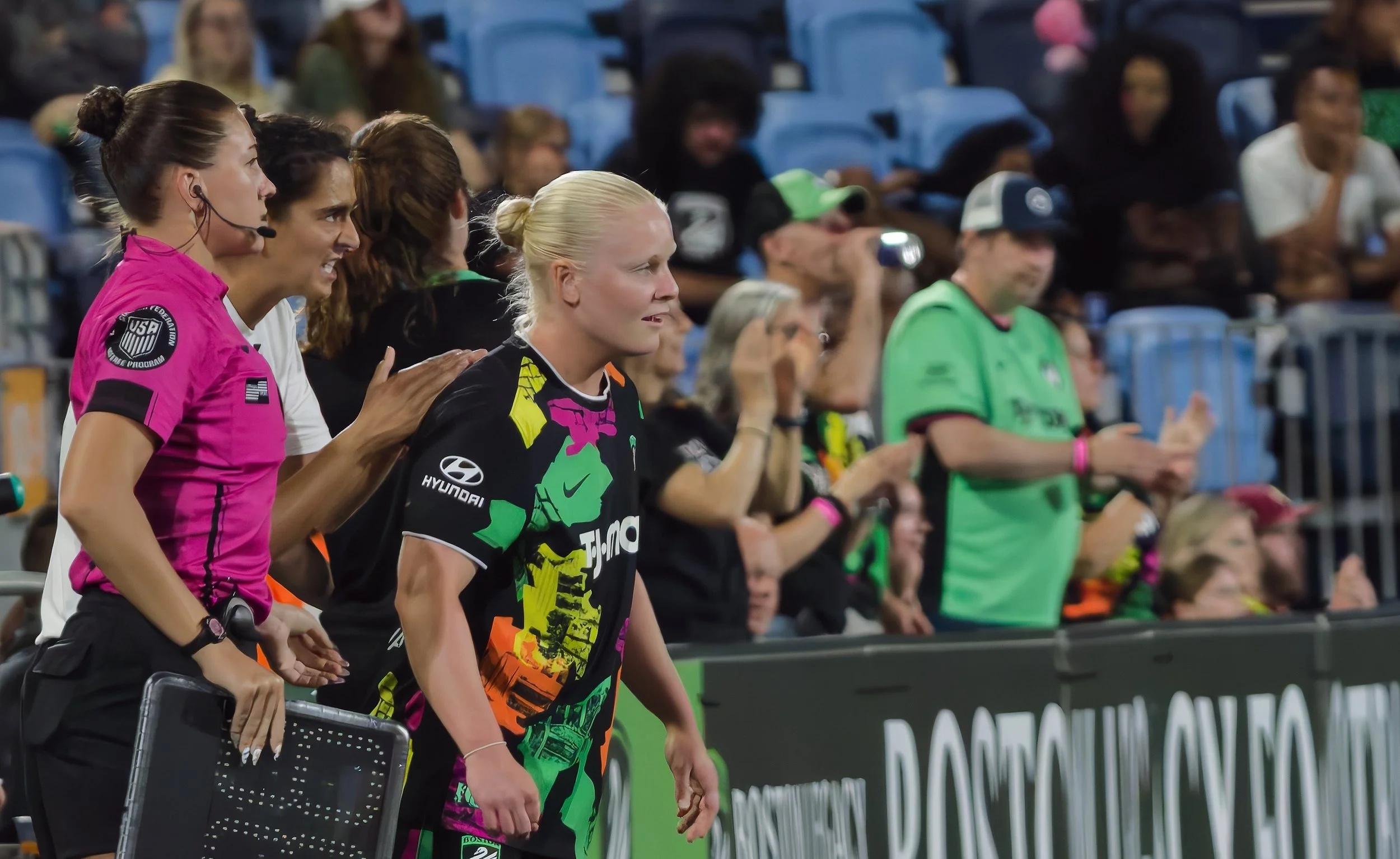 Women soccer players standing on the sidelines during a match, with spectators seated in the background.