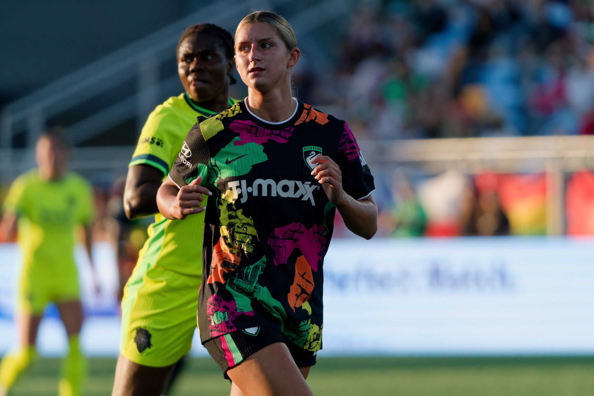 Two female soccer players running on the field, with stadium and crowd in the background. The player in the foreground wears a colorful black, pink, orange, green, and yellow jersey with the Boston logo and the Nike swoosh. The second player is sligh