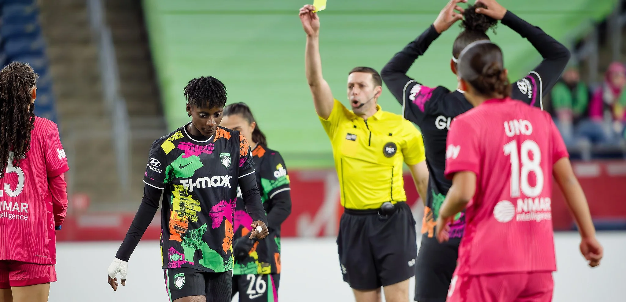 Soccer players and a referee on the field during a match, with players wearing pink and black jerseys, and the referee in a yellow shirt.