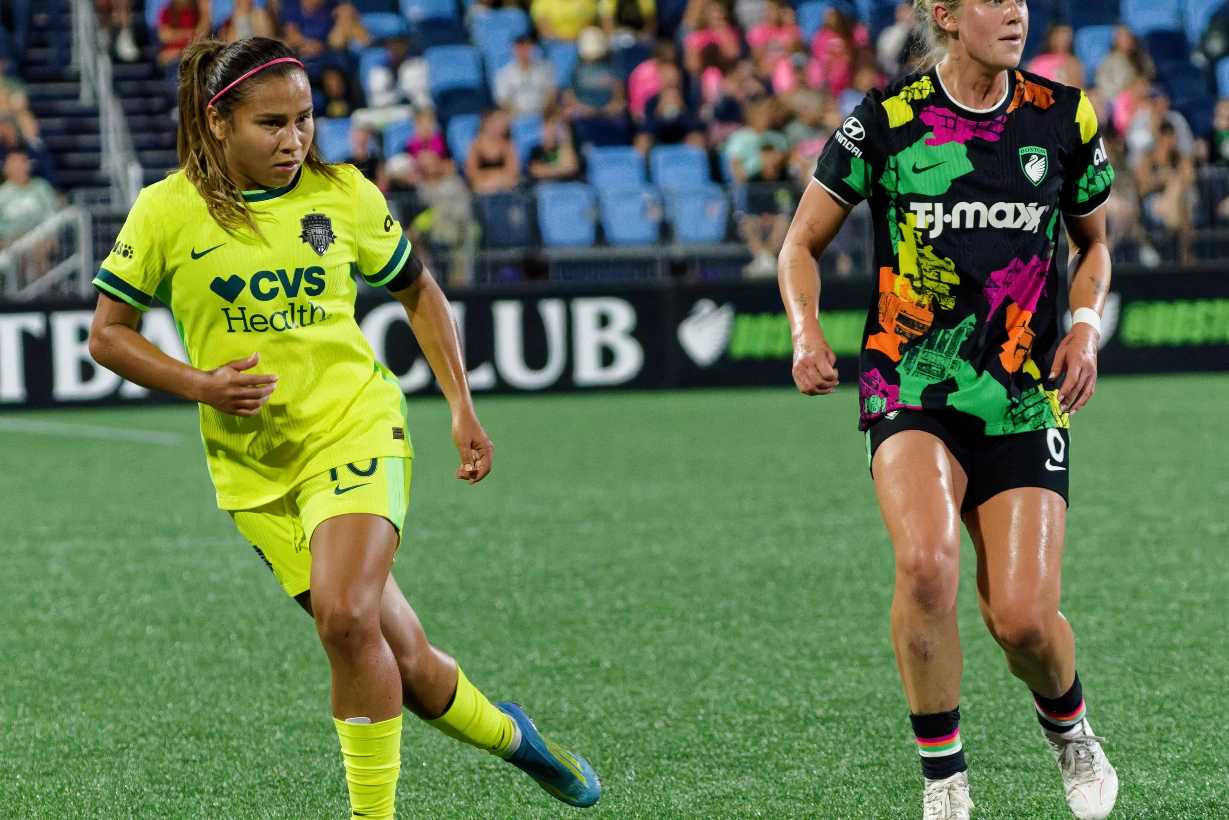 Two female soccer players on the field during a game, one in a bright yellow uniform and the other in a colorful black and green uniform. The background shows a blurred audience in stadium seats.