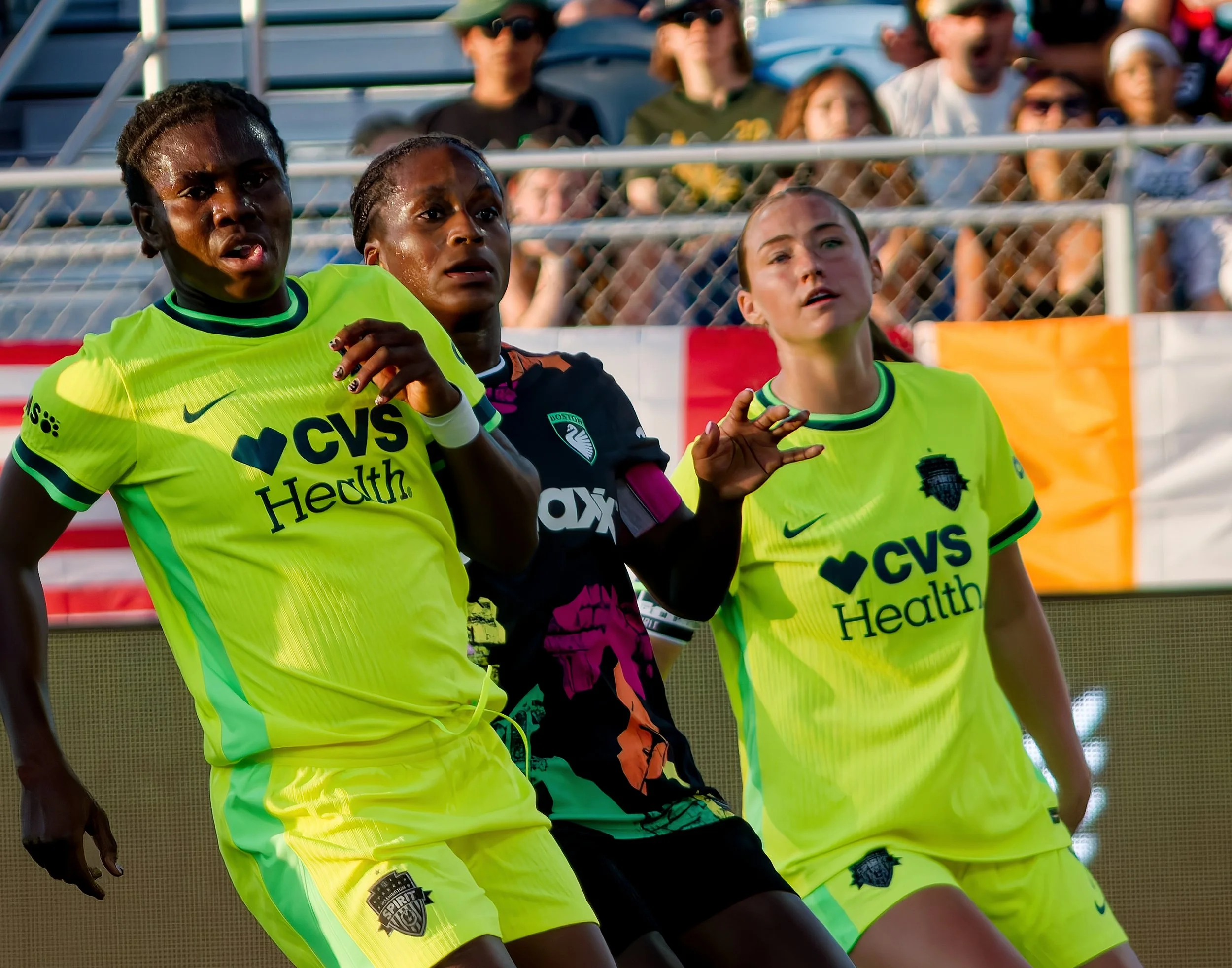 Three women in neon yellow sports jerseys with the logos "CVS Health" and "Spirit" on sports field, focused on the game, with spectators in background.