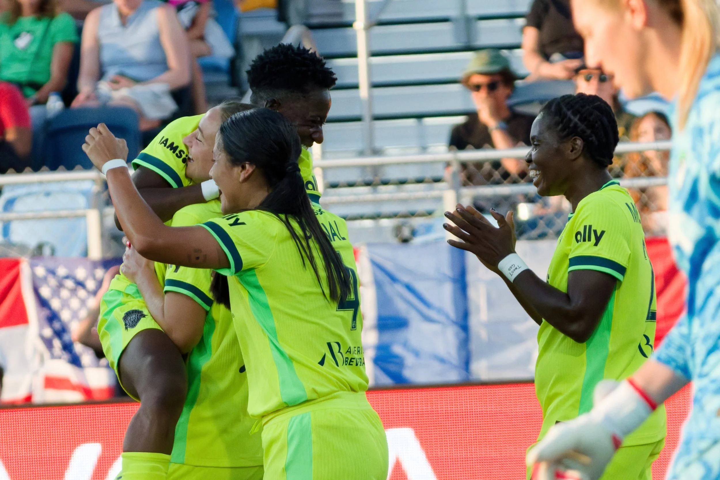Soccer players in bright yellow jerseys celebrating on the field, with one player hugging another, and fans in the stands behind them.