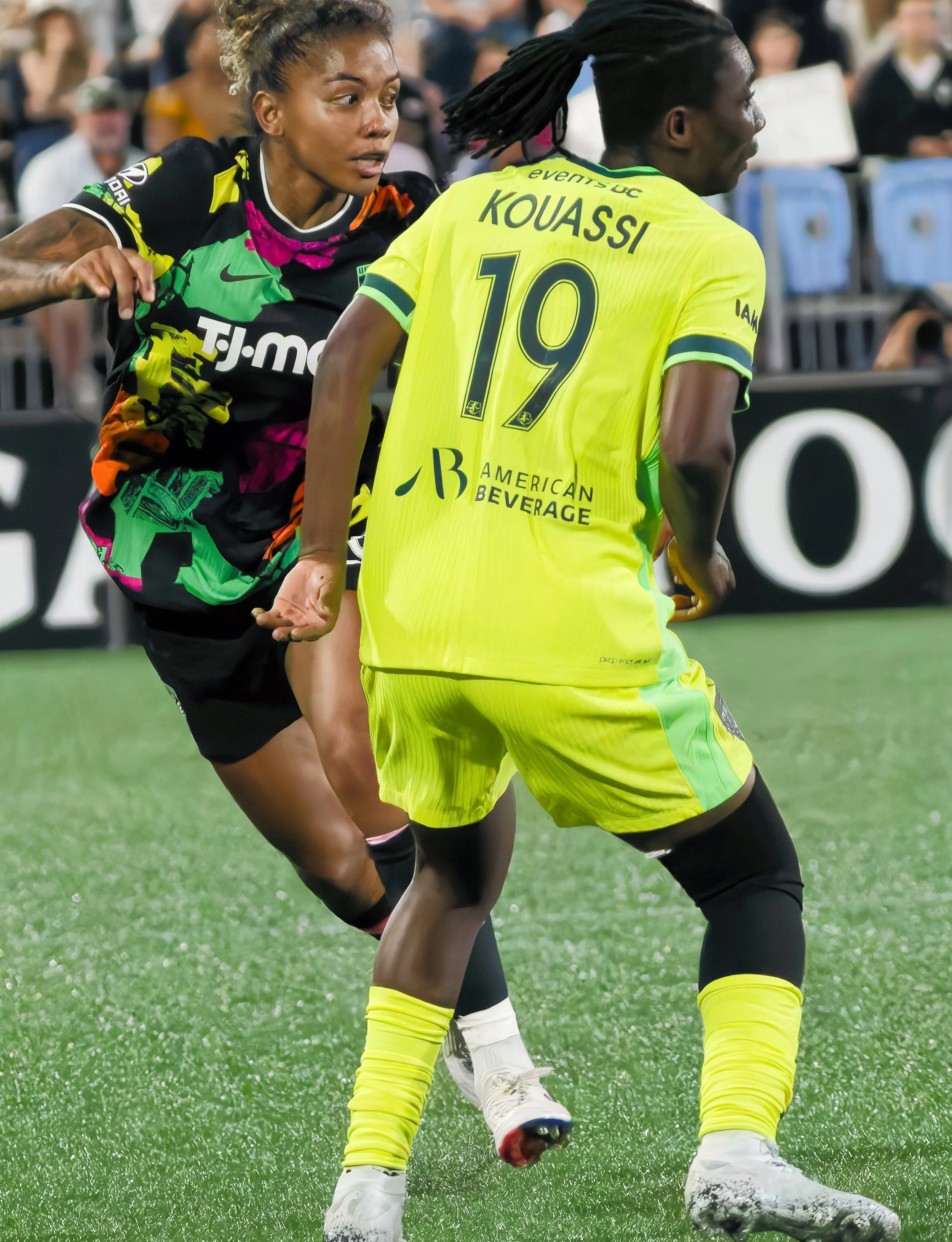 Two soccer players with dark skin compete for the ball on a field. One wears a green and black uniform, and the other is in a yellow uniform with 'KOUASSI' and the number 10 on the back. The game is in a stadium with a blurred crowd in the background