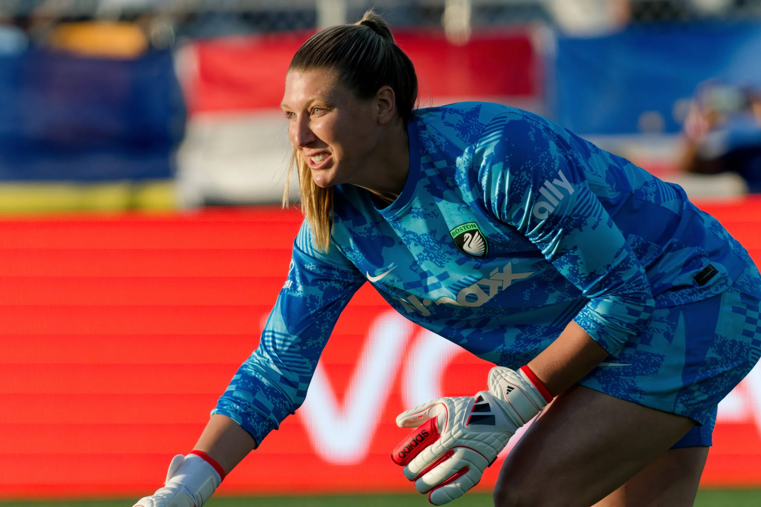 A female soccer player wearing a blue jersey and white Adidas gloves, crouching on the field during a game.