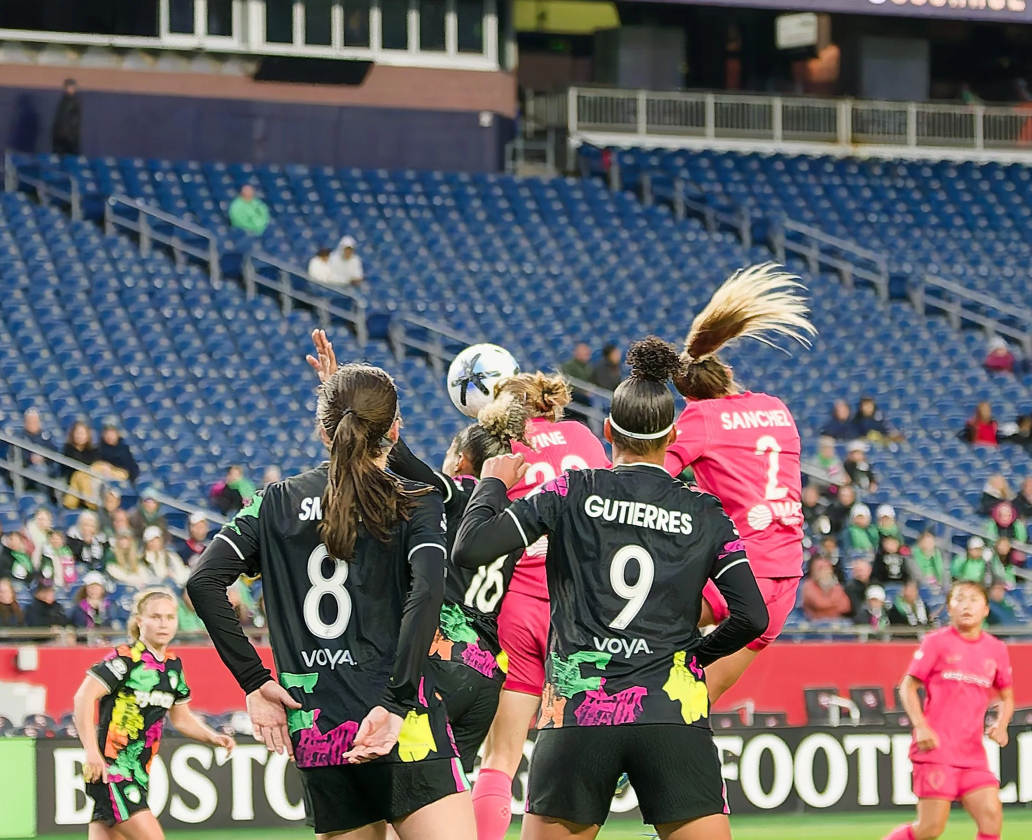 Women playing soccer, wearing black and pink jerseys, on a soccer field with a stadium and spectators in the background.