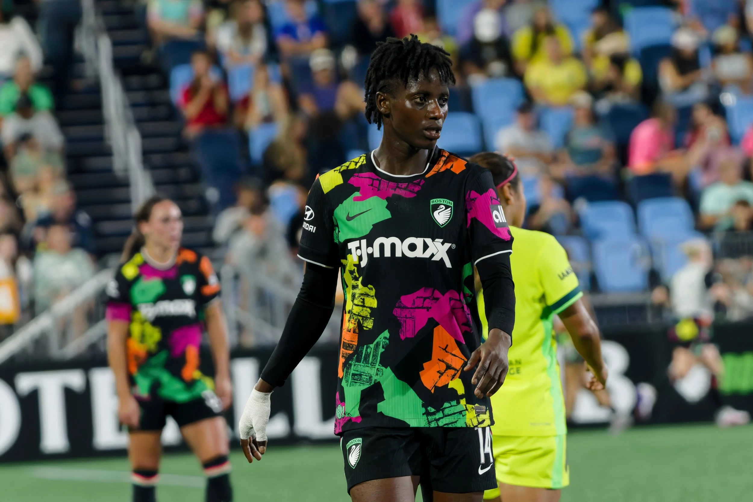 A female soccer player with short dreadlocks in a colorful uniform, standing on a soccer field with spectators in the background.