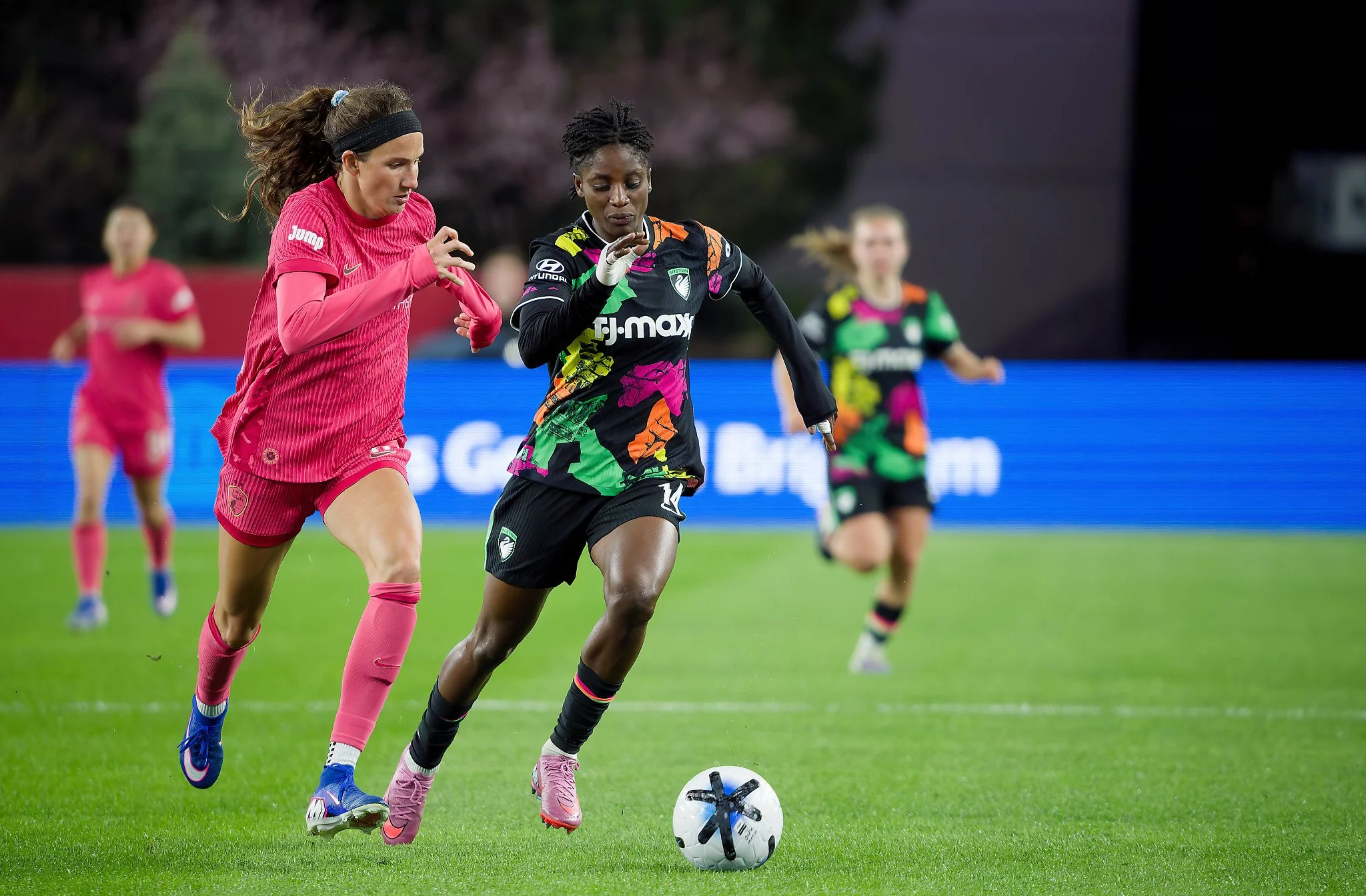 Two soccer players running after a soccer ball during a match, one in pink uniform and the other in black uniform with colorful patterns.