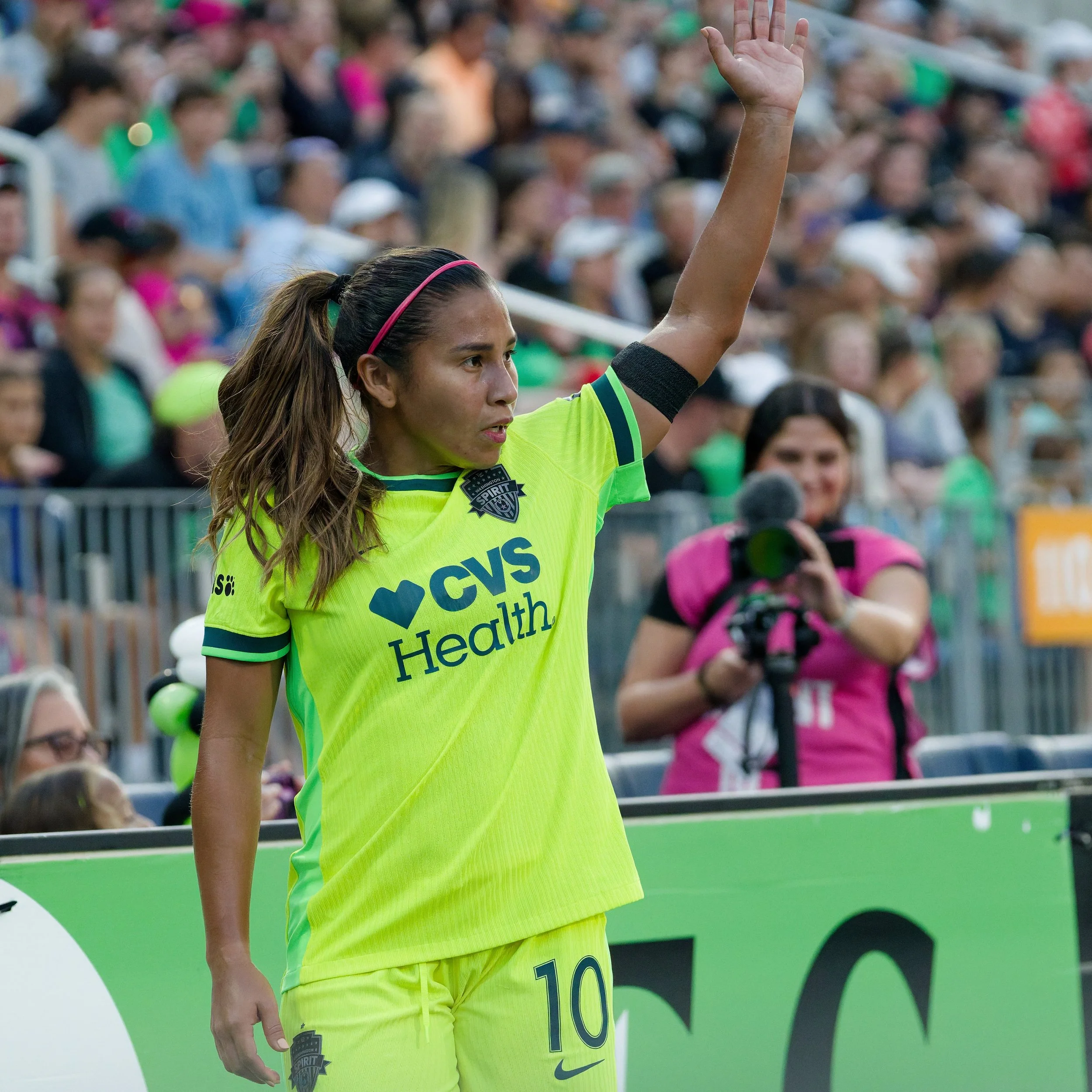 A female athlete in a yellow sports jersey raising her right arm in front of a blurred crowd at a sporting event.