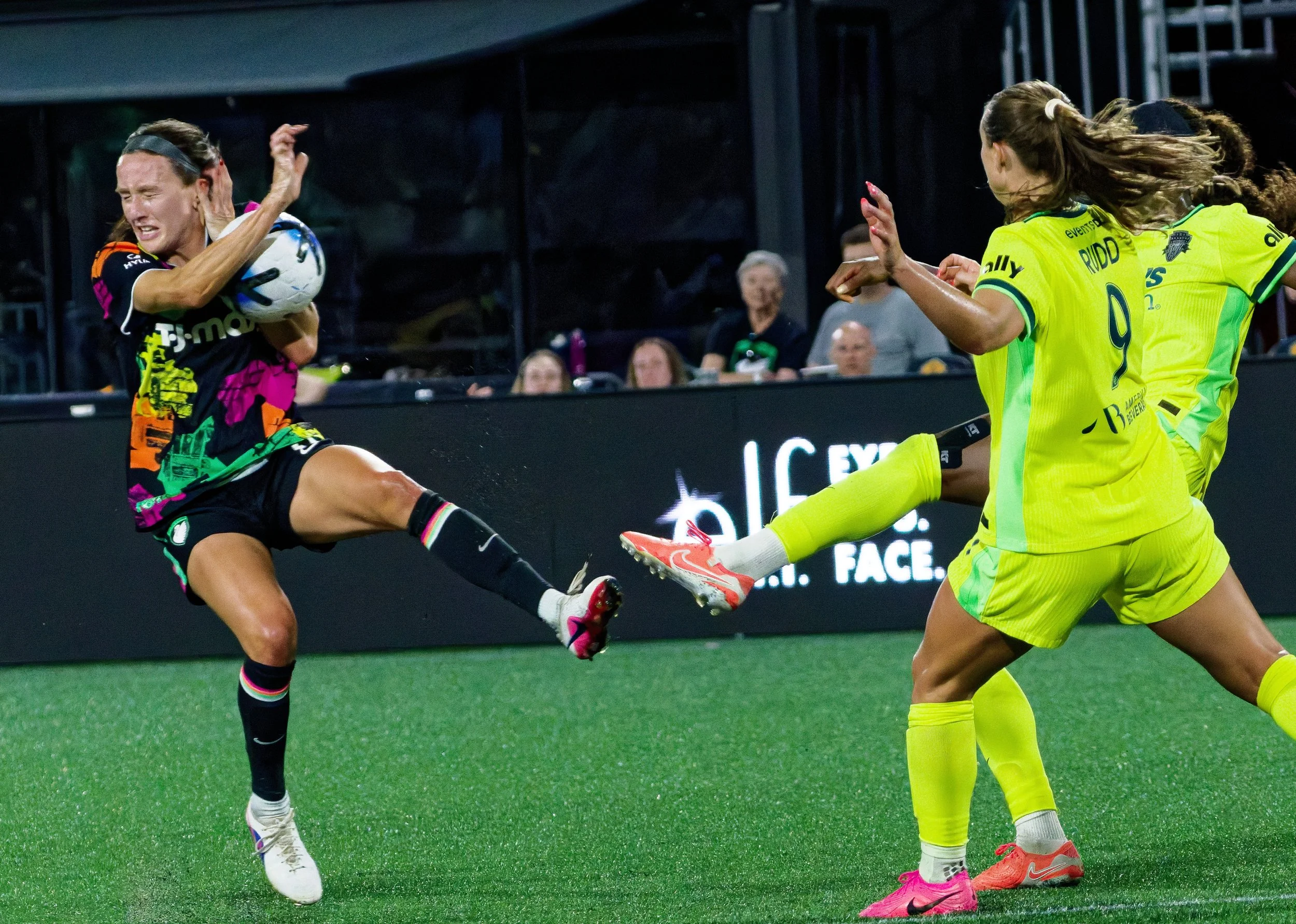 Two women playing soccer, one in a black and colorful jersey kicking the ball and the other in a yellow jersey blocking the kick, with spectators in the background.