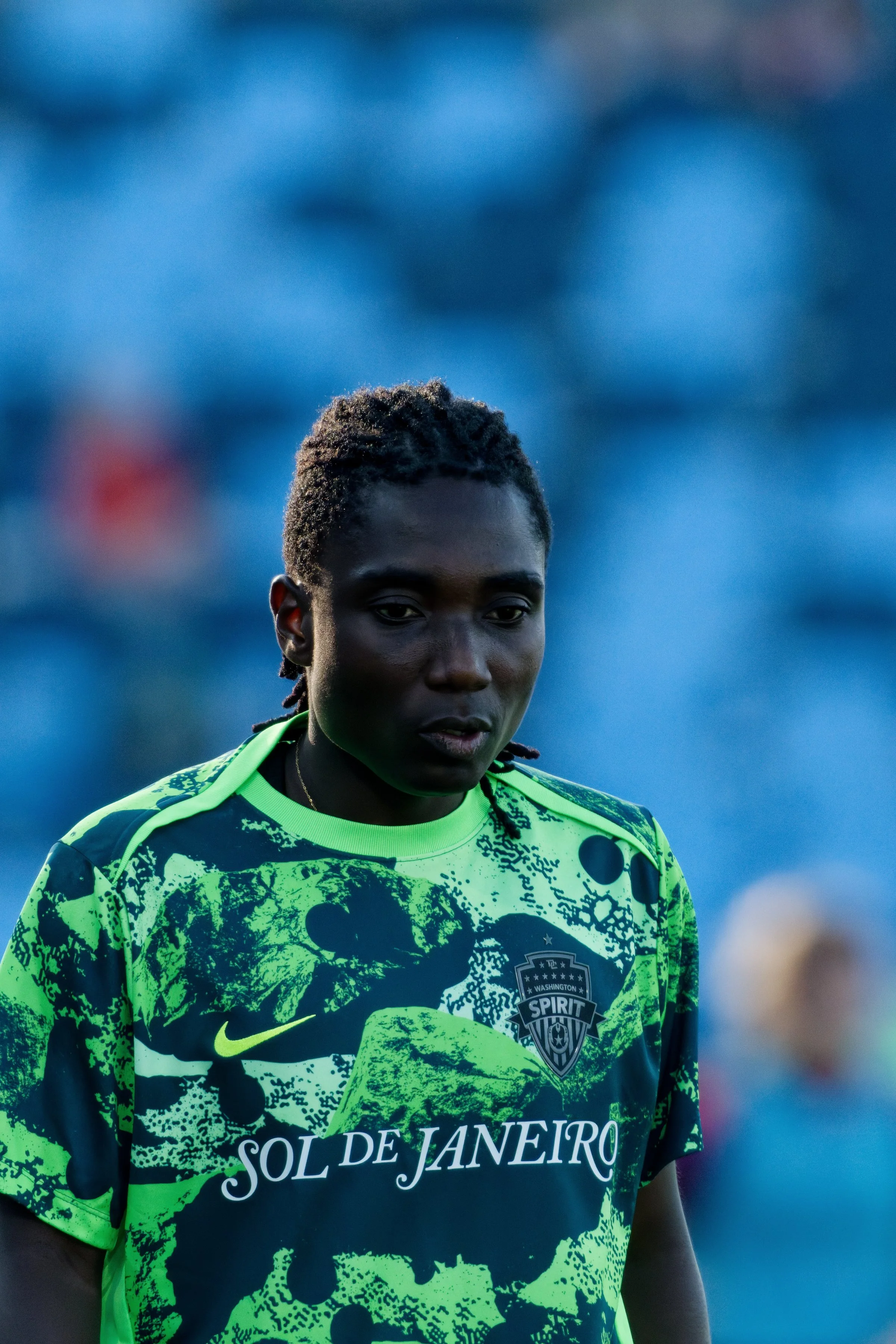 A male soccer player with dark skin and short curly hair, wearing a bright green and black sports jersey with the words 'Sol de Janeiro' and a badge that reads 'Washington Spirit'. He is outdoors with a blurred background.
