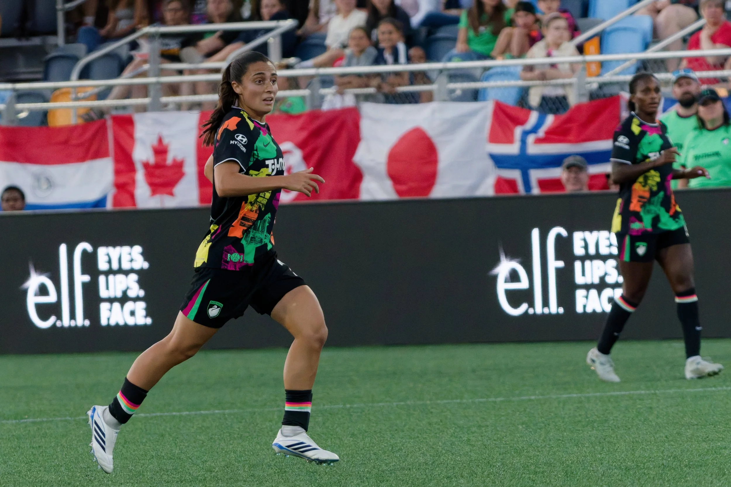 A female soccer player running on the field during a match, wearing a colorful black and green uniform with rainbow accents, with fans in the stands and Canadian flags in the background.
