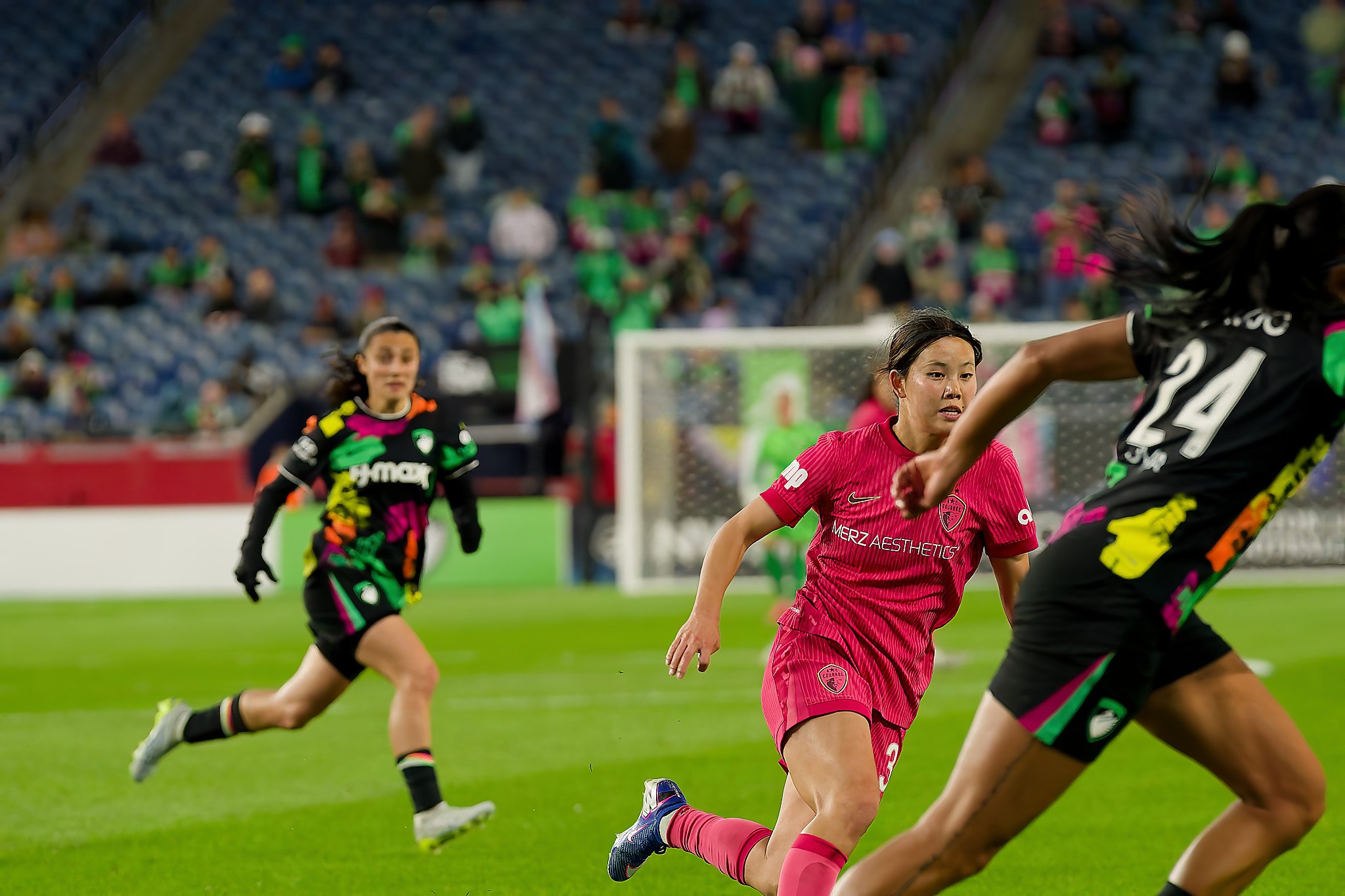 People playing soccer on a field in a stadium with a blurred crowd in the background.