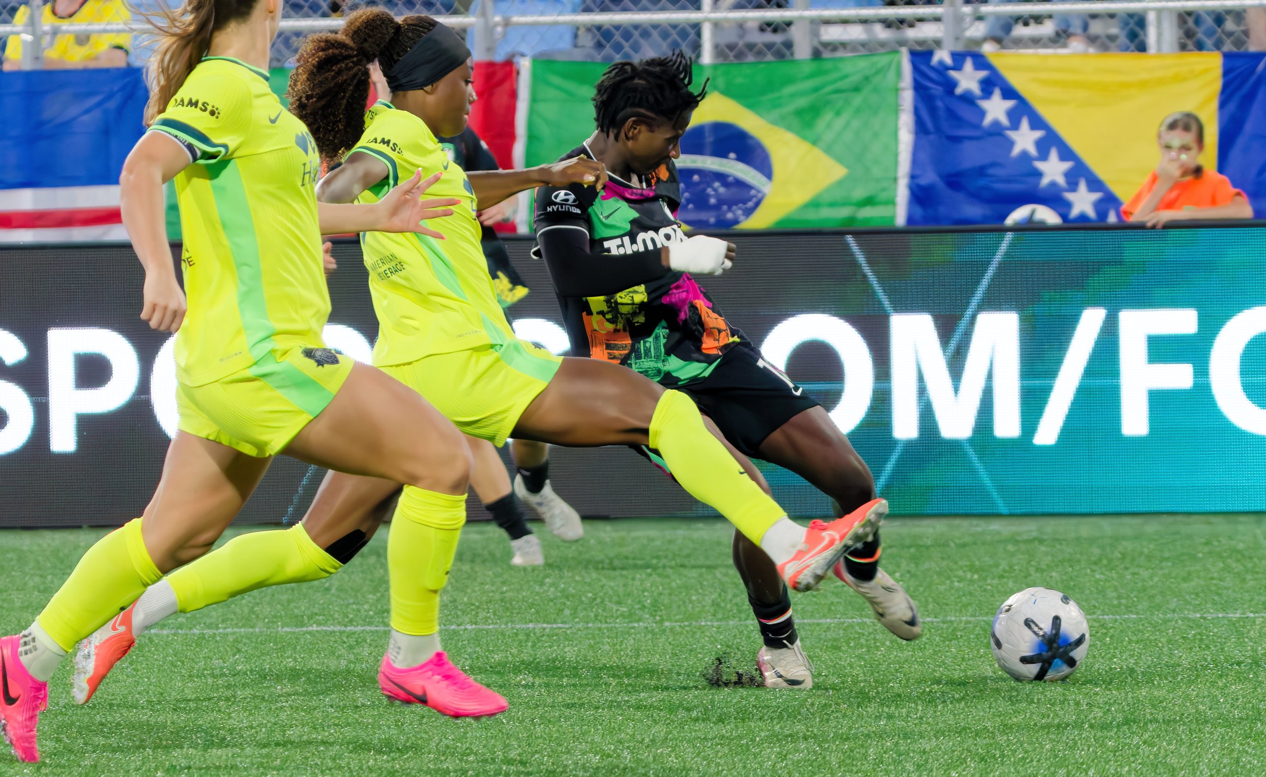 Women soccer players in action, competing for the ball on the field. Two players are wearing yellow uniforms, and one player is wearing a black and multicolored uniform. There are flags of Bosnia and Brazil in the background.