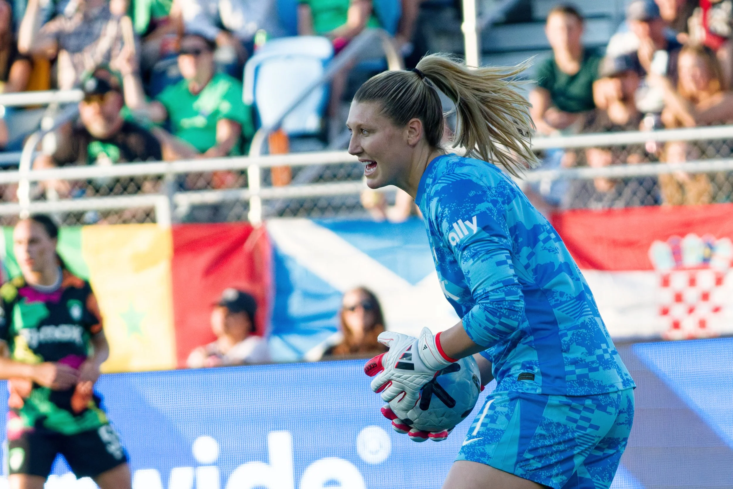 A female soccer goalkeeper in a blue uniform and gloves is holding a soccer ball during a match, with spectators in the background.
