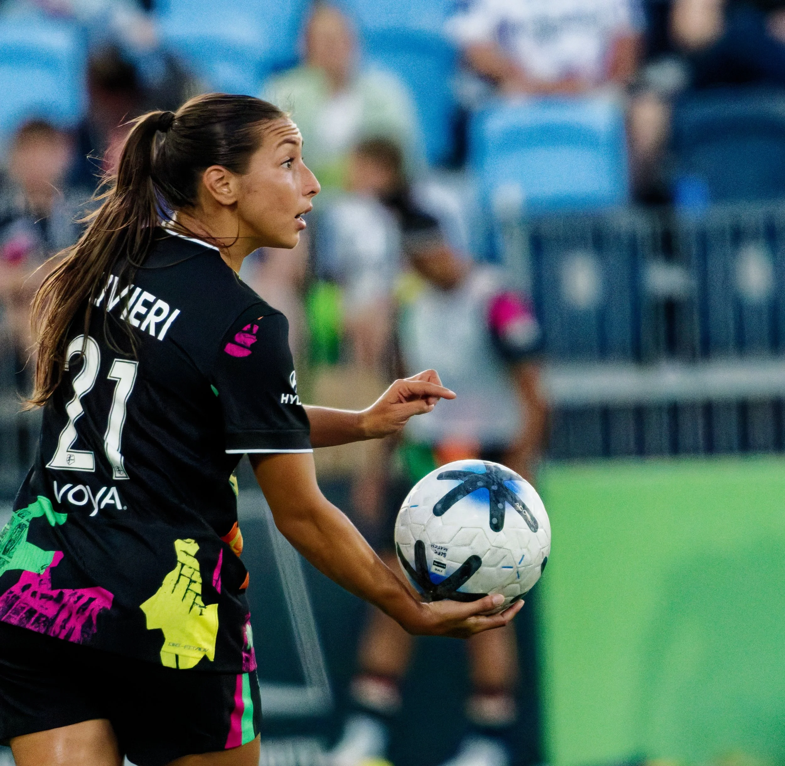 A female soccer player in a black and pink uniform holding a soccer ball during a game.