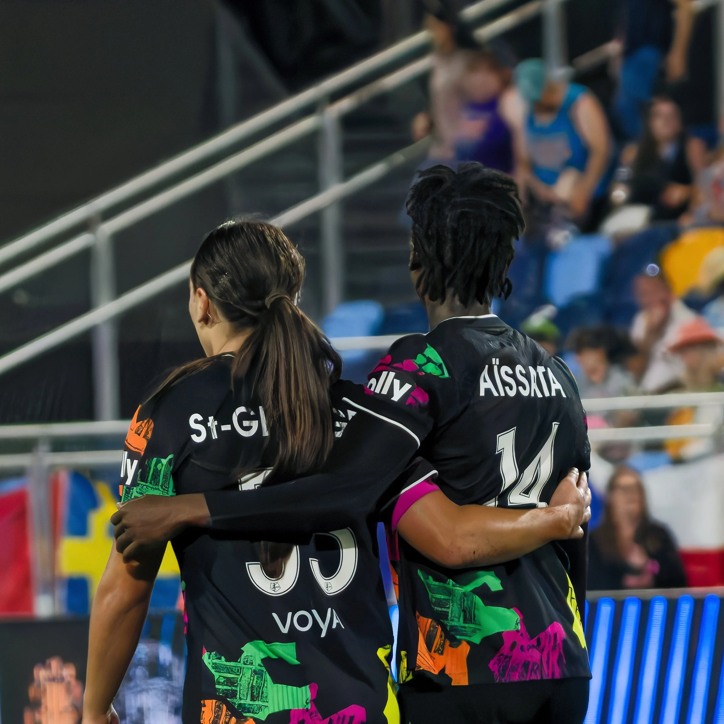 Two soccer players in black jerseys hugging during a game, with a group of spectators blurred in the background.