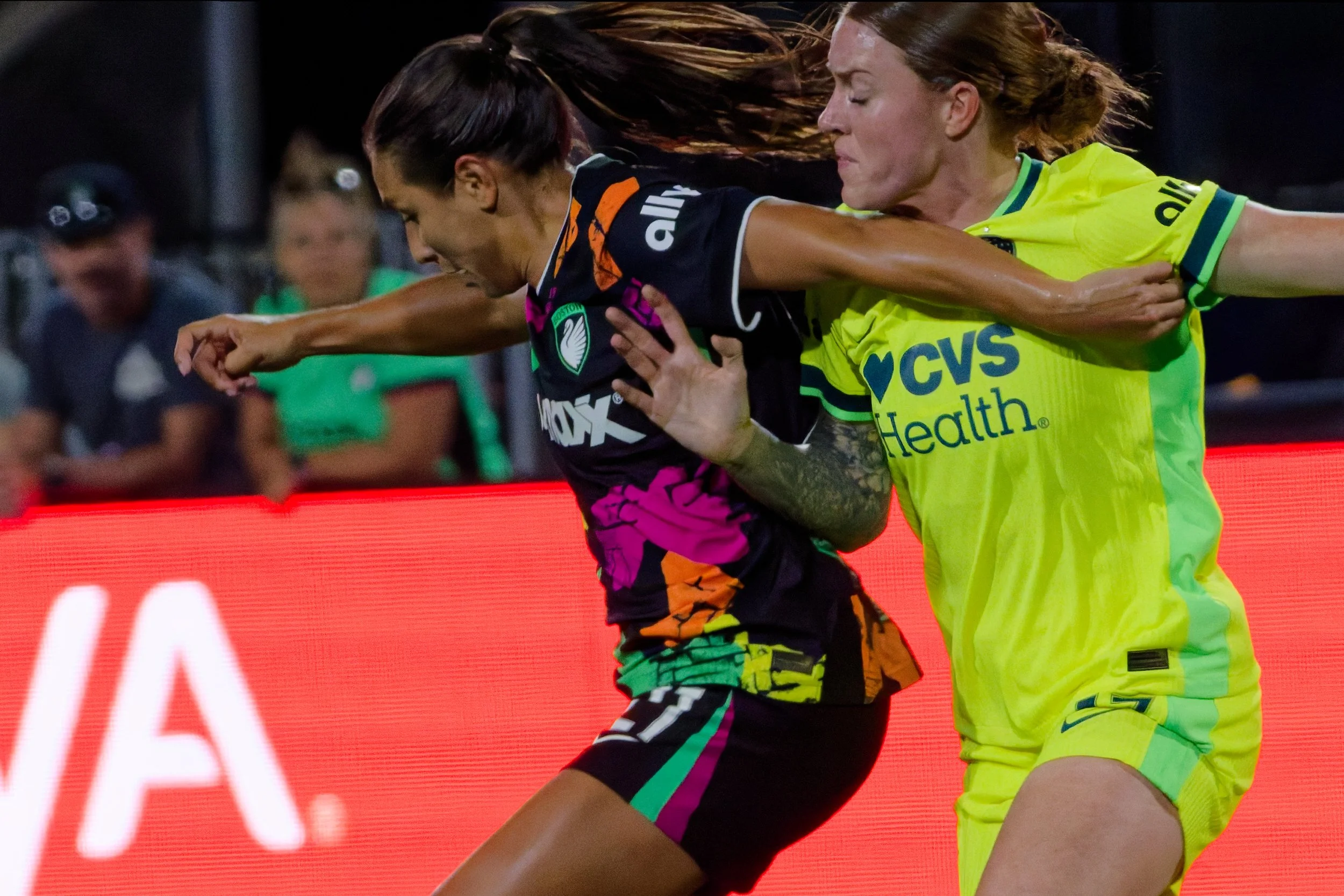 Two female soccer players engaged in a physical contest during a match, with spectators blurred in the background.