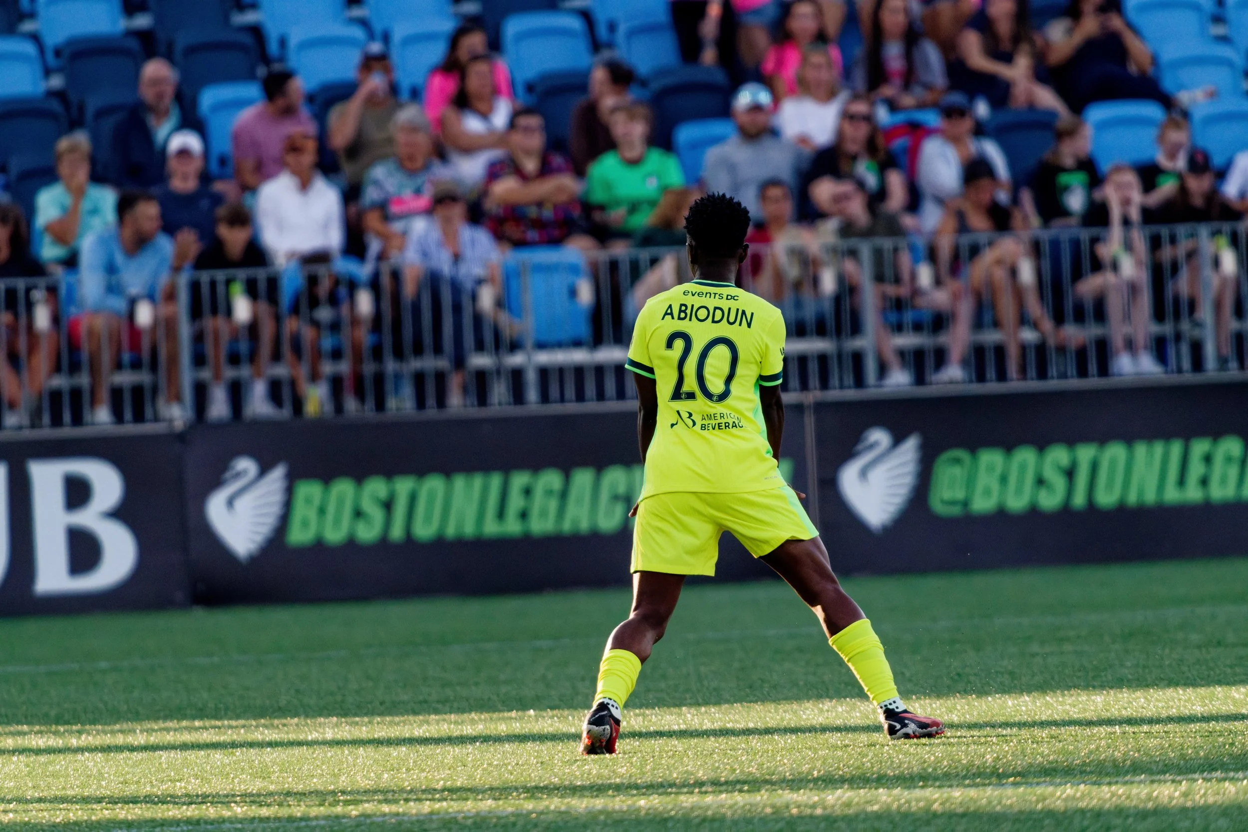 A soccer player wearing a yellow uniform with the name ABIDOUN and the number 20 on the back, standing on a soccer field during a game, with a crowd of spectators seated in blue stadium seats in the background.