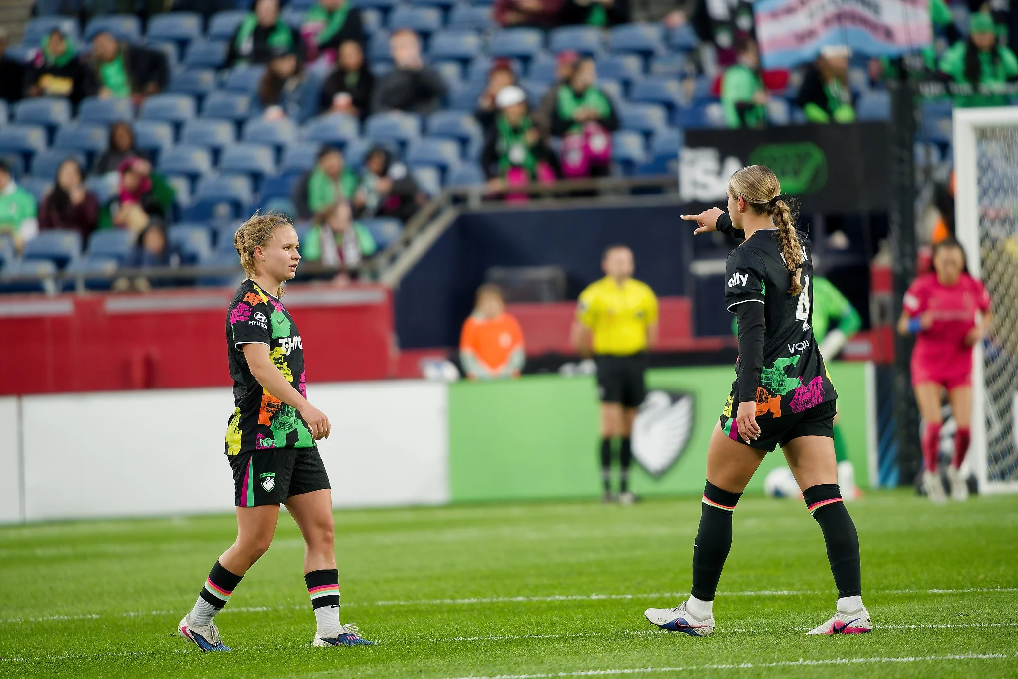 Two soccer players in black and colorful uniforms standing on a soccer field and communicating, with a crowd in green and black in the stands in the background.