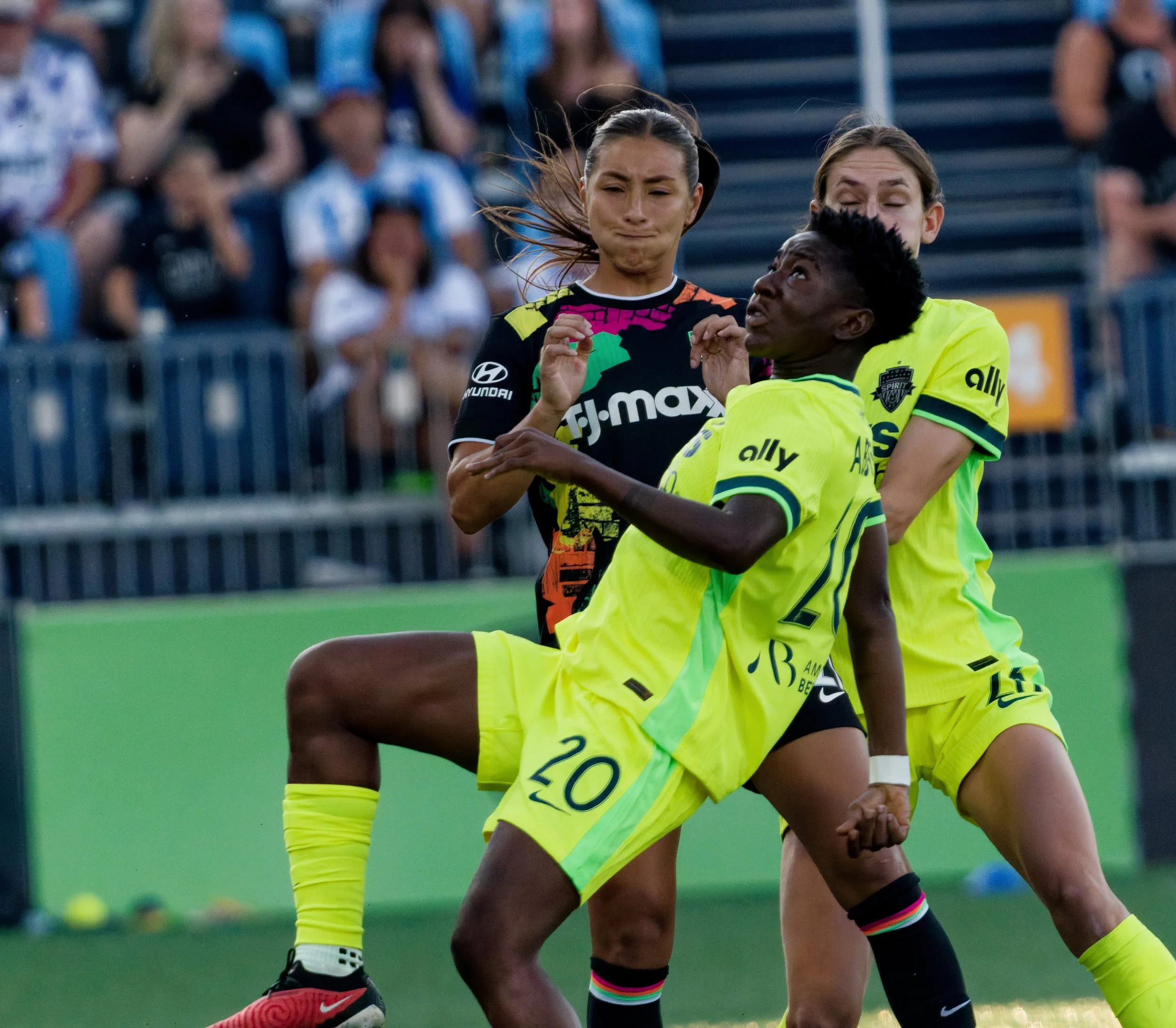 Two female soccer players in yellow and black uniforms in a physical contest during a game, with spectators in the blurred background.