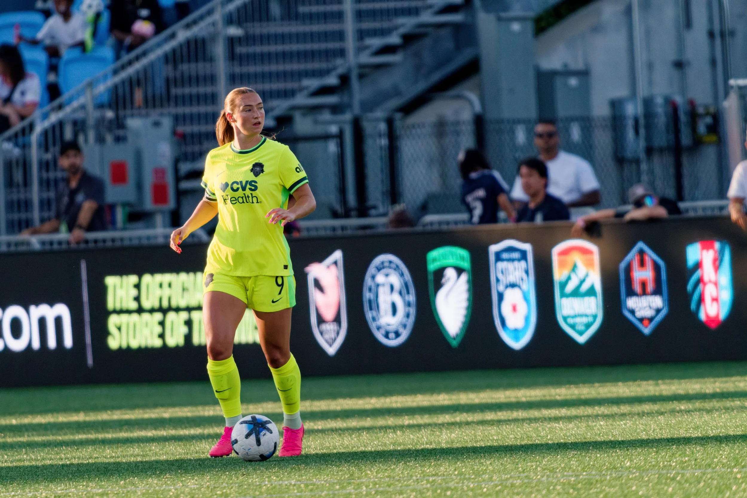 A female soccer player in a neon yellow uniform with the number nine, standing on a soccer field with a ball at her feet. She has long hair tied back and is wearing pink cleats. There are fans in the stands behind her, with some sitting and some stan