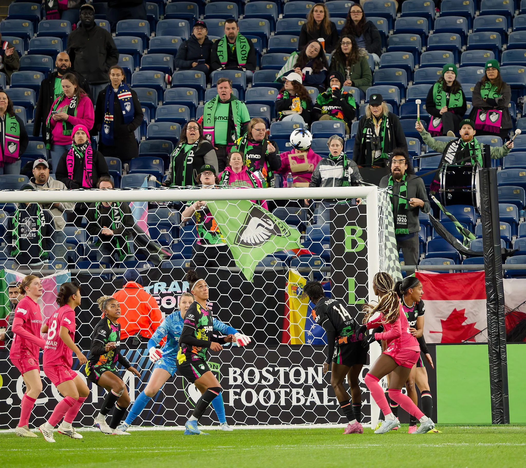 soccer players on the field during a match, some wearing pink jerseys and others in multicolored uniforms, with fans in the stands behind the goal.