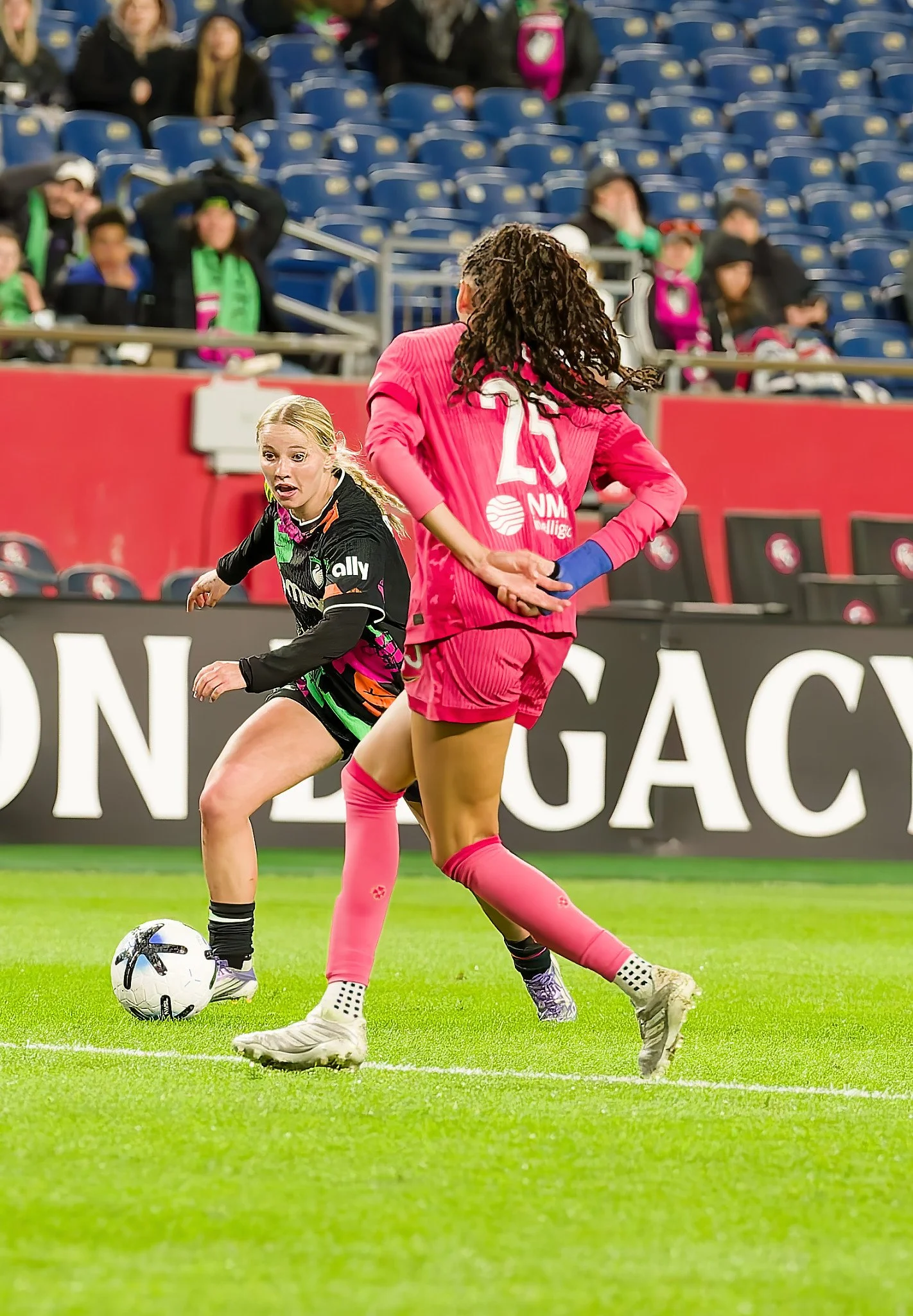 Two soccer players competing for possession of the ball on the field, one in a black and pink uniform and the other in a pink uniform, with spectators in the background.
