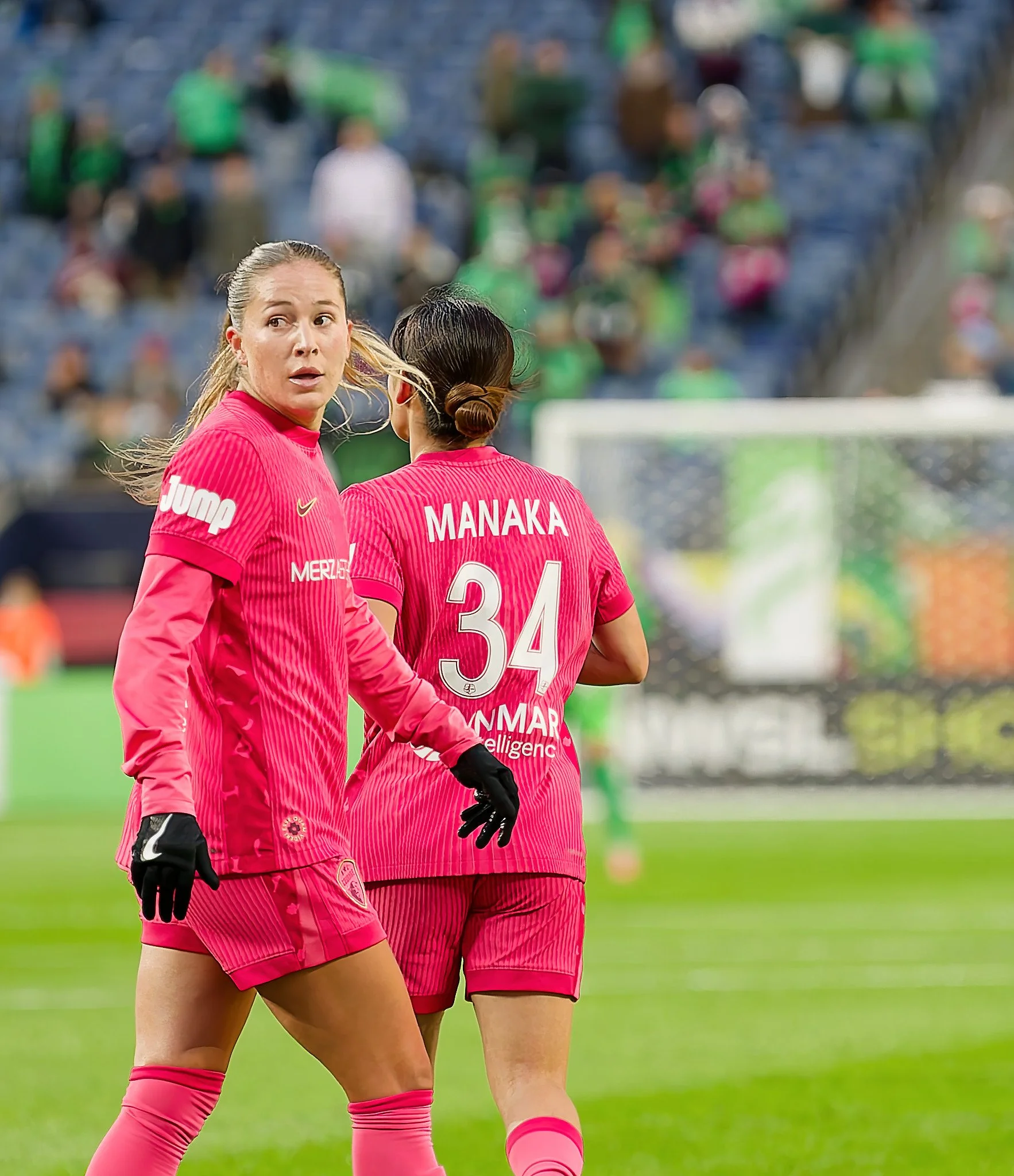 Two soccer players standing on the field, wearing pink uniforms. One has the name 'MANAKA' and the number '34' on her back. The standoff is outdoors in a stadium with a blurred crowd in the background.