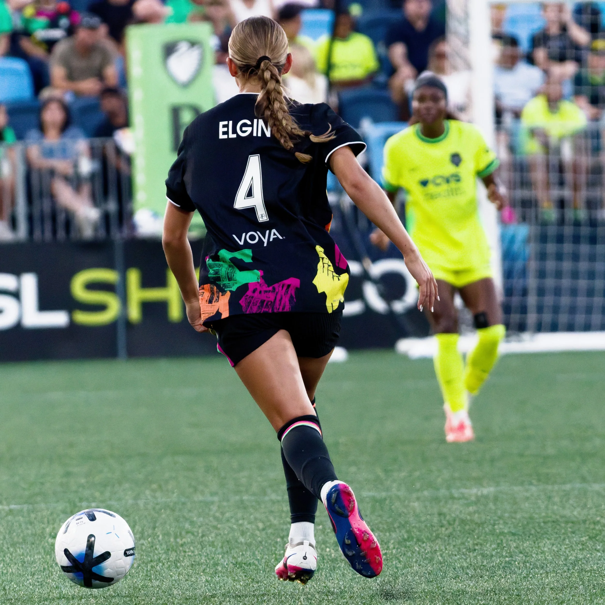 A female soccer player with a ponytail, wearing a black jersey with the name 'Elgin' and the number 4, is kicking a soccer ball on a green field during a game. Other players and spectators are visible in the background.