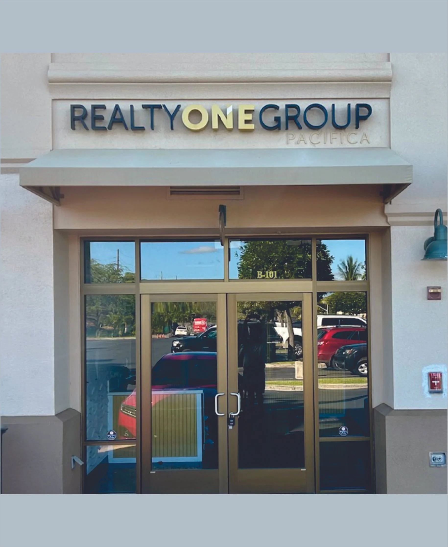 The entrance to a real estate office named Realty One Group Pacifica, with glass doors, a sign above, and a reflection of parked cars and trees in the glass.