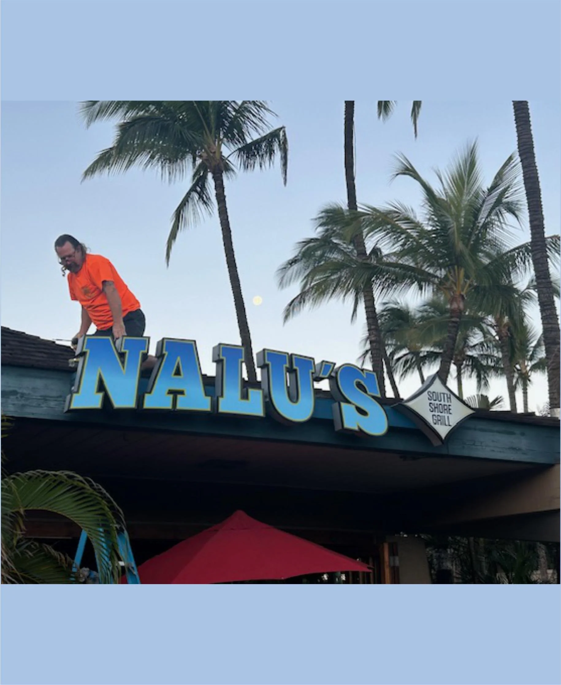 A person installing or repairing a sign that reads 'Nalu's', with palm trees and a clear sky in the background, Maui Hawaii.