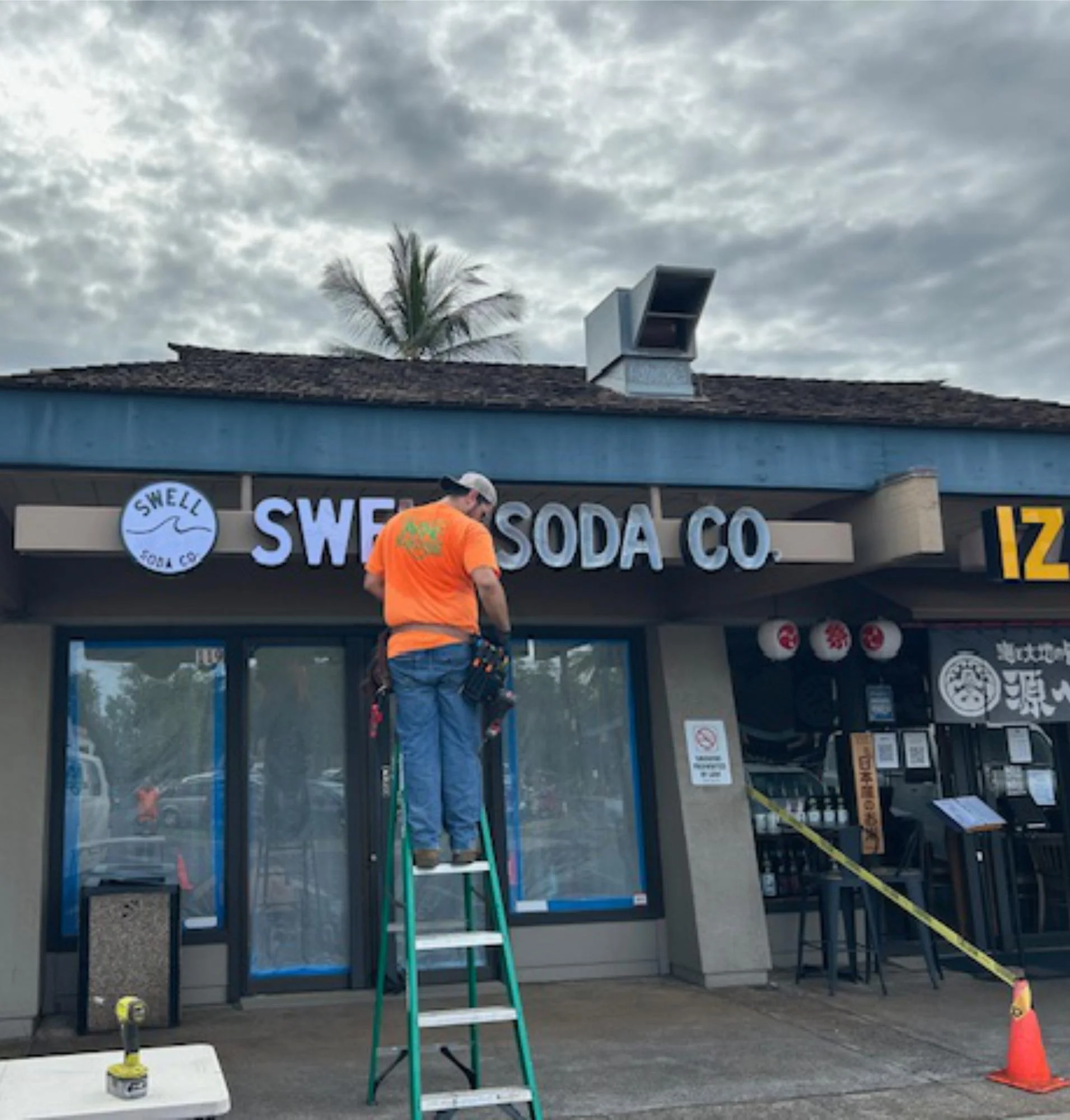 A worker on a ladder installing a sign that reads 'SWELL SODA CO' in front of a building with a palm tree in the background. The sky is cloudy and there are construction cones and equipment around him.