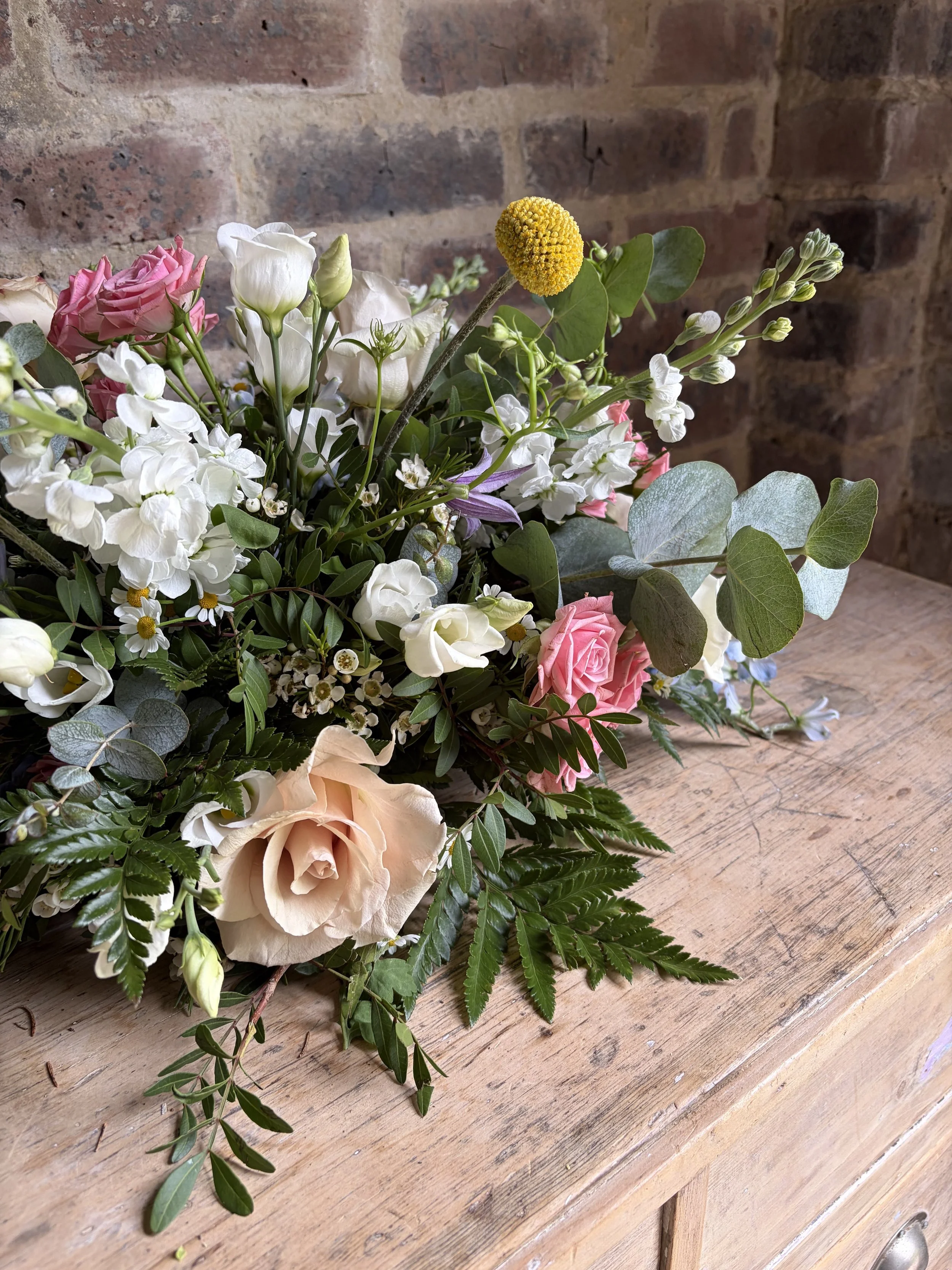 A bouquet of mixed flowers, including pink roses, white lisianthus, yellow billy buttons, and various greenery, resting on a wooden surface with a brick wall in the background.