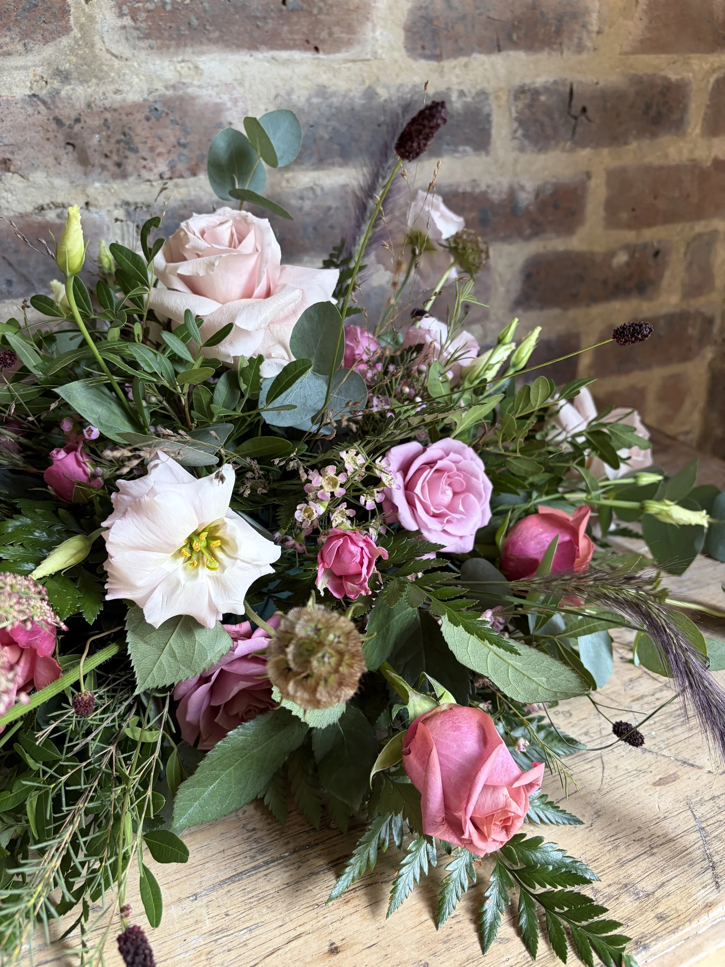 Close-up of a floral arrangement with pink roses, green leaves, and various other small flowers, placed on a wooden surface with a brick wall in the background.